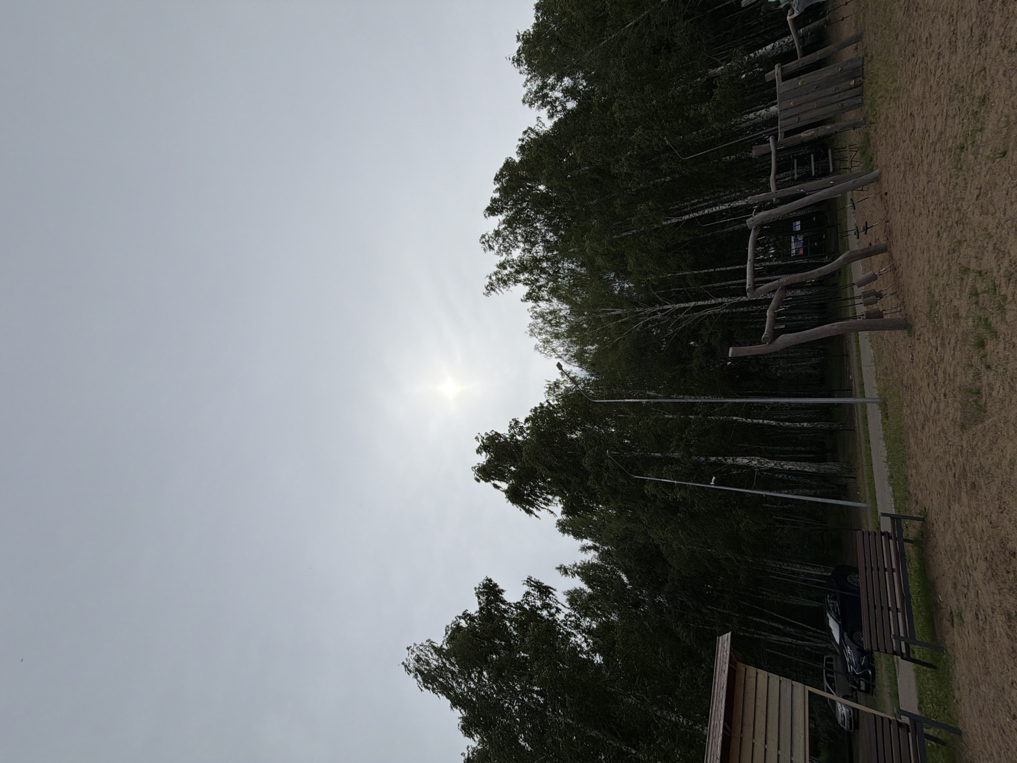 The image depicts a park area with a sandy ground, a playground structure made of natural wood, and several trees, primarily birches, lining the background. Two wooden benches are visible in the foreground, and a small wooden shelter or gazebo is located to the left. Cars are parked near a path that runs along the edge of the trees. The sky is overcast with the sun faintly visible as a bright spot. This scene is located in Chaikovsky, Russia.