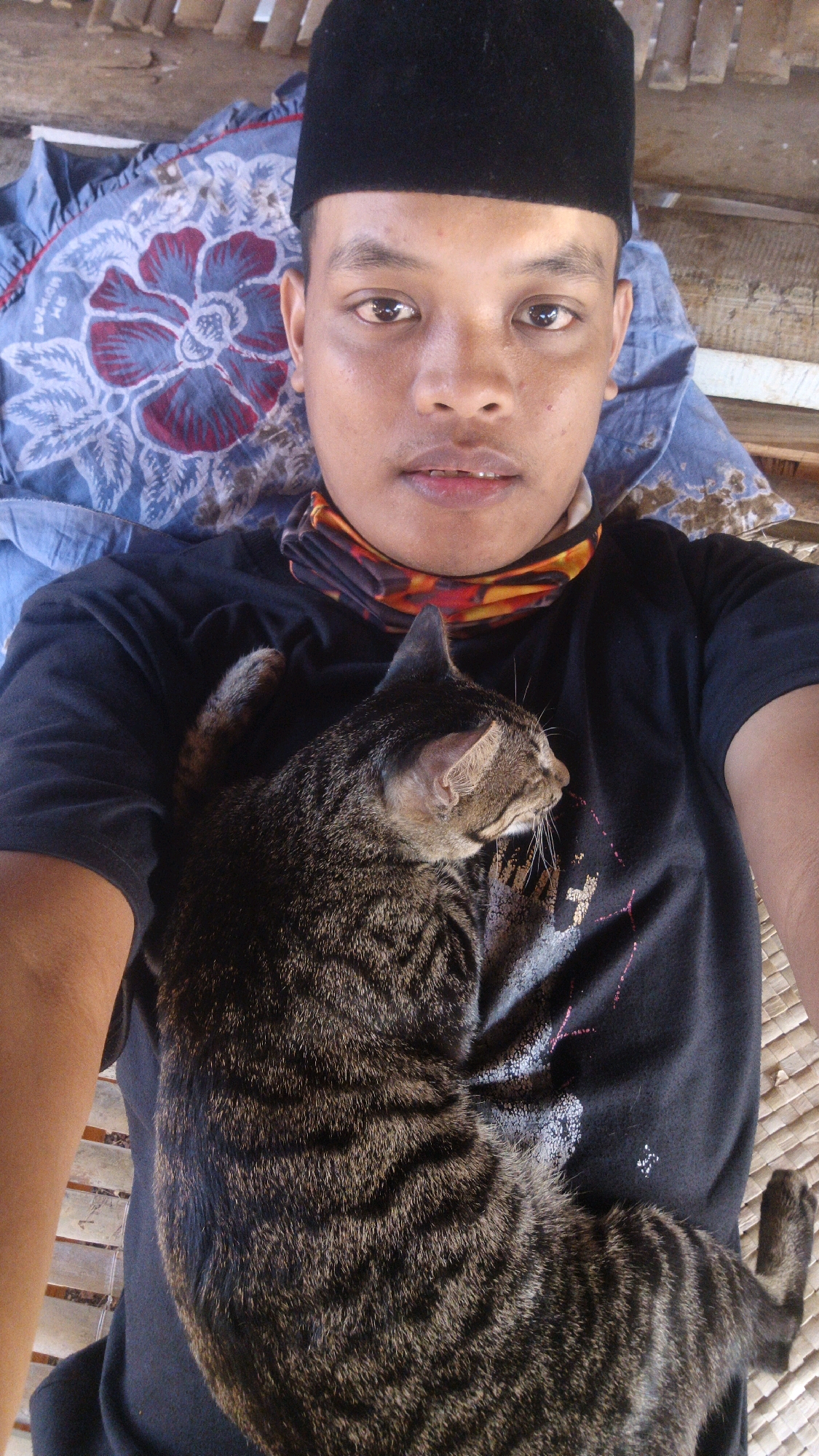 A person is lying down outdoors with a cat on their chest. The person is wearing a black t-shirt, a black cap, and a multi-colored bandana around their neck. The cat is a tabby with dark stripes. The background appears to be a woven mat and bamboo. The person is taking a selfie, as evidenced by their face being directed towards the camera. The location is Sokaramme Timur, Indonesia.
