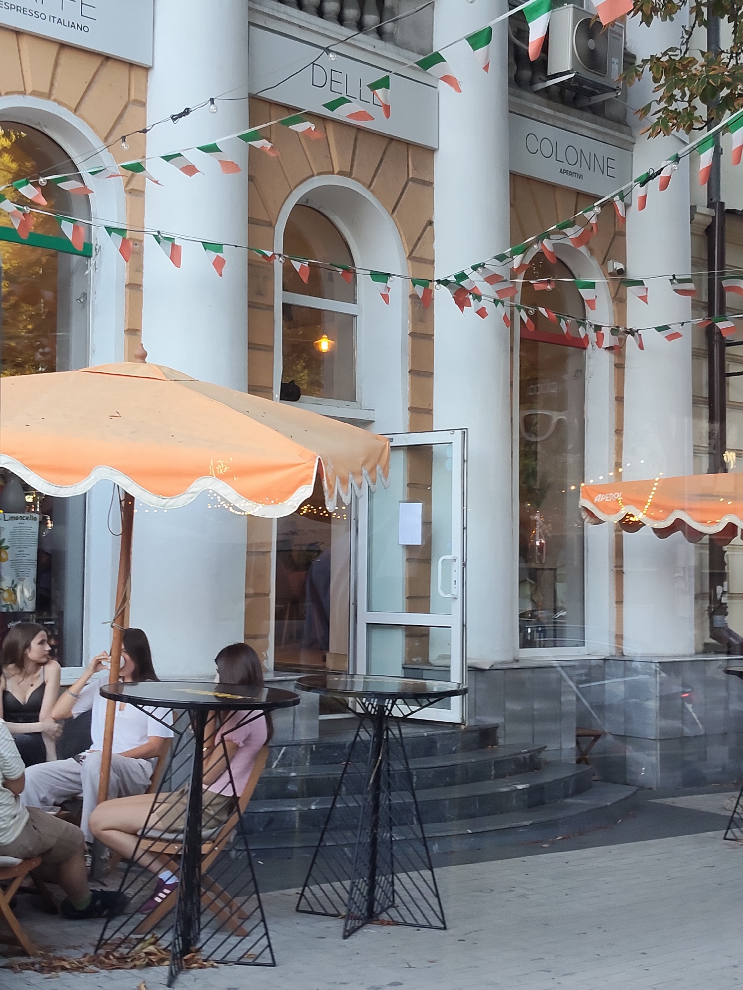 The image depicts an outdoor seating area of a cafe or restaurant, likely located in Chișinău, Moldova, given the prominent Italian flag decorations. Several people are seated at tables under an orange umbrella. The building facade features large arched windows and white columns. String lights are visible, as are decorative Italian flags. The ground is paved, and there are steps leading up to the entrance.