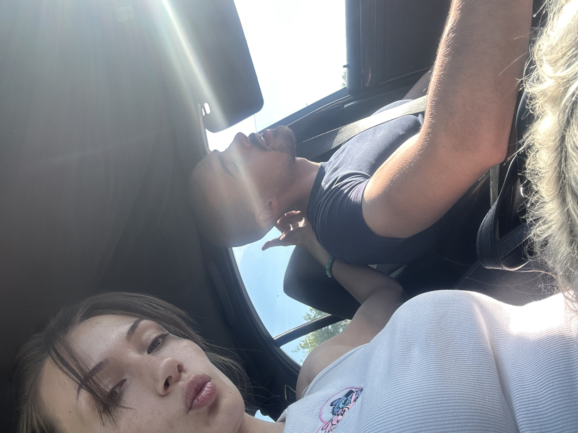 A selfie, presumably taken from inside a car, features two individuals. One individual, a woman, is positioned in the lower left corner, with a portion of her face and torso visible. She is wearing a white tank top with a cartoon graphic. The second individual, a man, is positioned above her, reclined in a seat and wearing a dark t-shirt. He is also wearing a seatbelt and has a small beard. Sunlight streams in through the car's sunroof, creating bright highlights. The location, Burgas, Bulgaria, cannot be determined from the image itself. No specific landmarks or distinguishing environmental details are visible. The interaction is a selfie, but no other direct actions are seen.