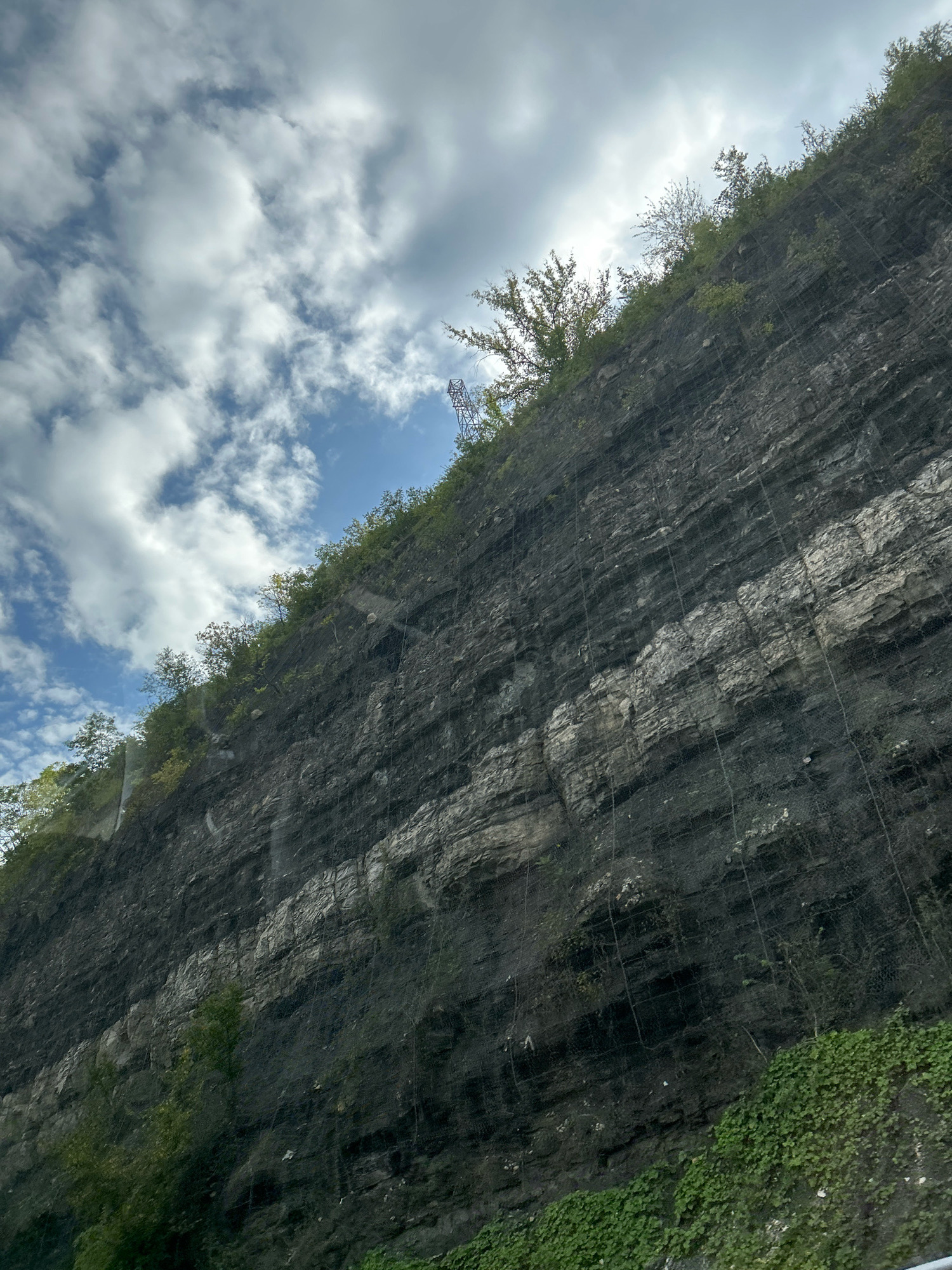 The image depicts a steeply sloped rock face covered in a dark, layered geological formation, characteristic of a quarry or a natural cliff face. The rock is largely dark gray with a distinctive horizontal band of lighter gray rock running through it, suggesting distinct geological strata. A fine, dark mesh netting is stretched across the face of the rock, extending from top to bottom, likely for erosion control or to prevent rockfalls. Sparse green vegetation, including small trees and ground cover, is visible growing on top of the rock face and at its base. The sky above is partly cloudy, with large, fluffy white clouds against a pale blue backdrop. The perspective suggests the image was taken from below and to the side of the rock face, possibly from a road or vehicle. The scene is devoid of any people or specific man-made structures aside from the netting. The location is identified as Collier Township, United States of America.
