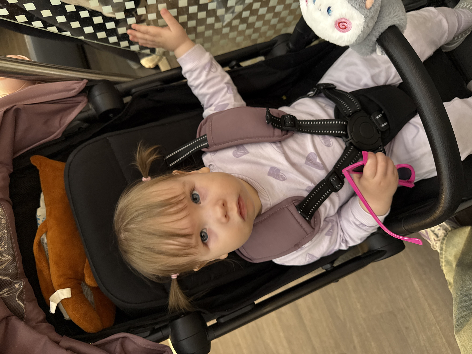 A young child with blonde hair in pigtails is secured in a black stroller. The child wears a light purple long-sleeve shirt and a harness. The child is holding pink glasses. A stuffed animal hangs from the stroller's handle. A brown stuffed toy is visible beside the child. The scene is indoors, possibly a home or retail establishment. The location is unspecified, and the claim of being in Moscow, Russia is unsubstantiated.
