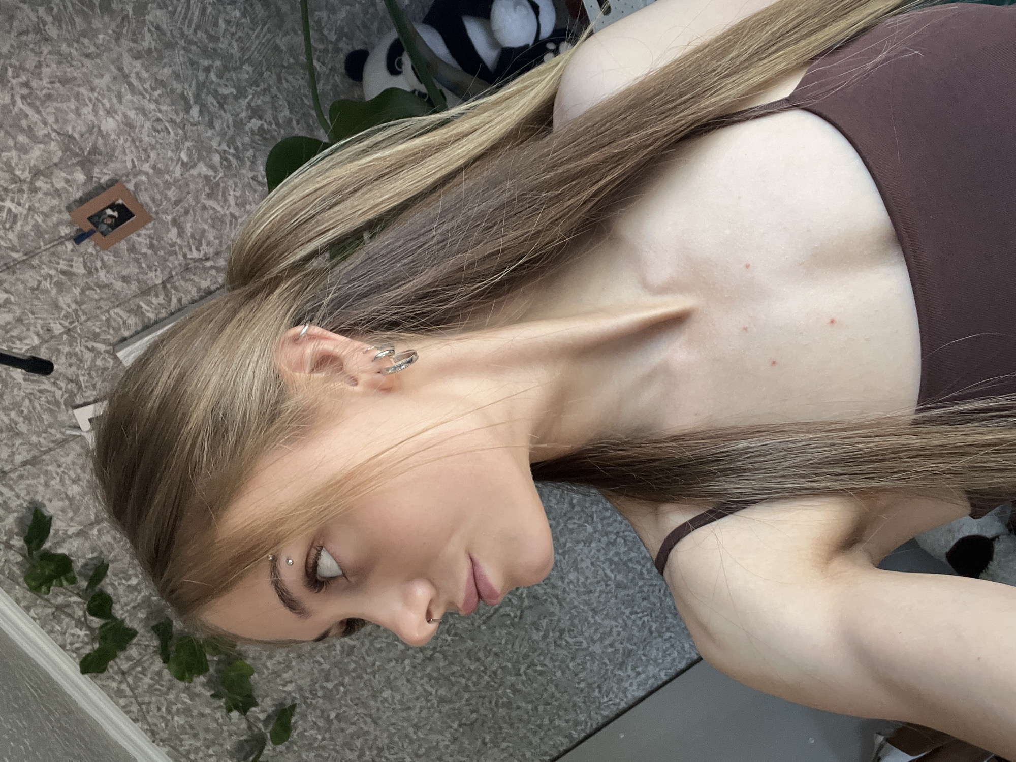 The image shows a close-up, profile view of a young woman with long, blonde hair tied in a ponytail. She has a nose ring and eyebrow piercing. She is wearing a dark brown tank top. The background appears to be an indoor setting with a textured wall, a plant with green leaves, and a stuffed panda toy visible. There are no discernable elements in the image that would allow for the determination of its location within the city of Фоки, Russia.