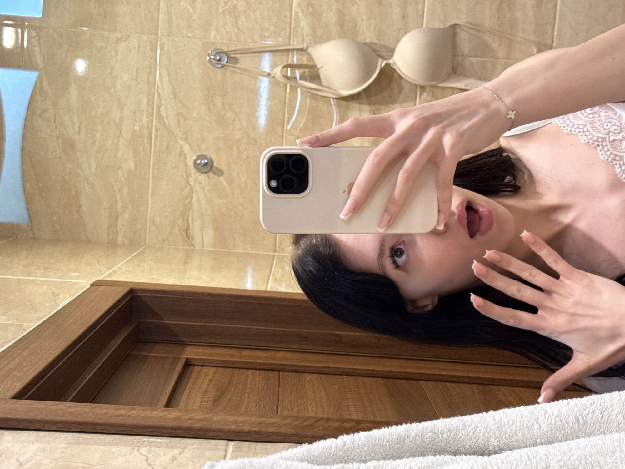 A young woman with dark hair is taking a mirror selfie in a bathroom. She is wearing a white camisole with lace trim and a delicate bracelet on her left wrist. Her right hand is holding up her phone, a pale pink iPhone, to take the picture. Her left hand is raised near her face, showing off her manicured nails. Behind her, a beige bra is hanging on hooks on a tiled wall. To her left, a white towel hangs on a wooden door. The bathroom walls are tiled with a marble-like pattern, and the door appears to be made of dark wood. The scene suggests a private, domestic setting, likely a home bathroom. The location provided, Khimki, Russia, is a city in Moscow Oblast.