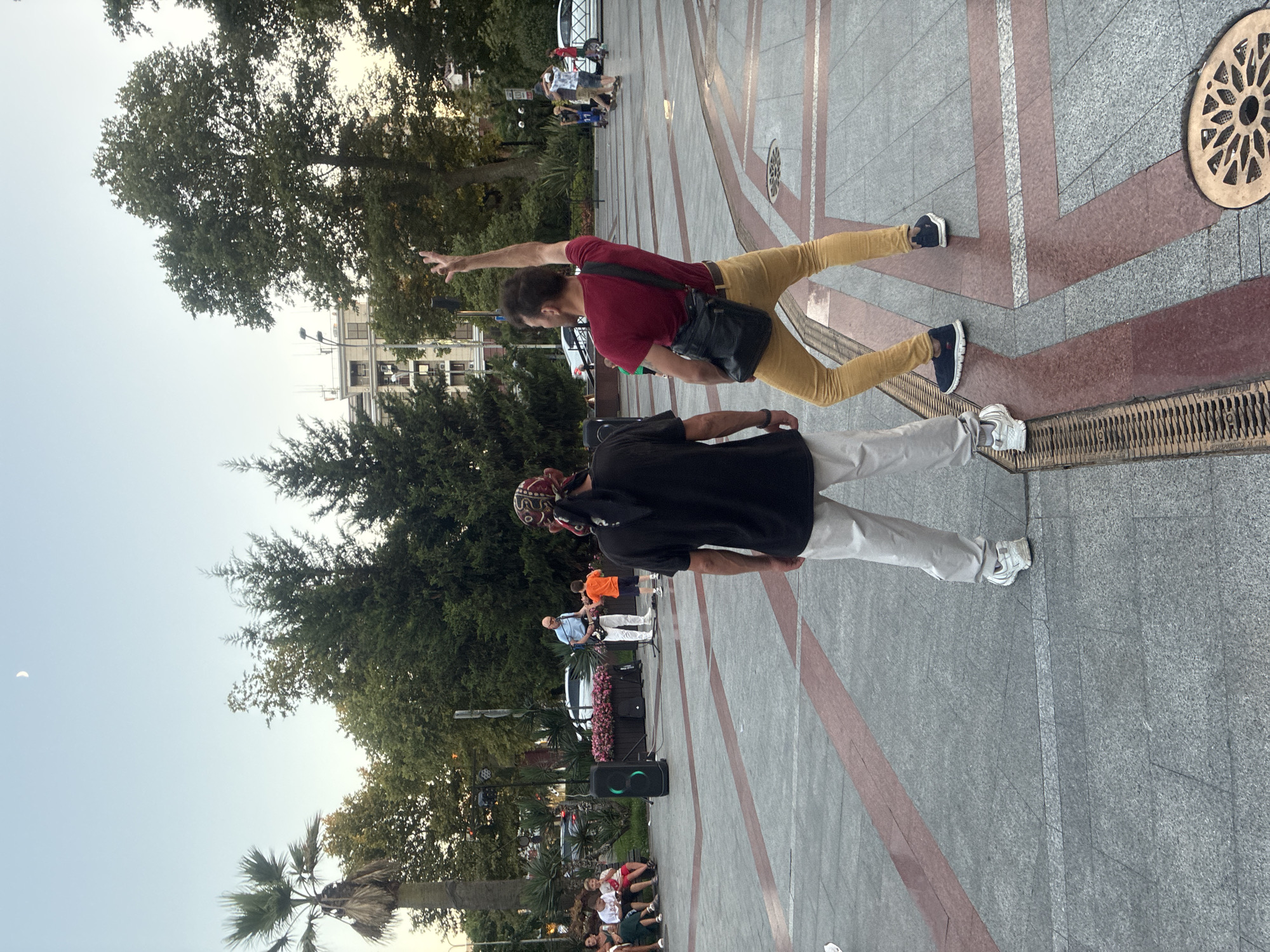 Two men are standing on a paved public square in Sochi, Russia, facing away from the camera. The man on the left is wearing a black t-shirt, white pants, and a patterned bandana on his head. The man on the right is wearing a red t-shirt, yellow pants, and has a black bag slung over his shoulder; he is gesturing with his right hand. In the background, a man is playing a guitar and singing, with a young boy standing next to him. Several other people are seated and standing around the square, and trees and buildings are visible behind them. The sky is clear with a sliver of the moon visible.
