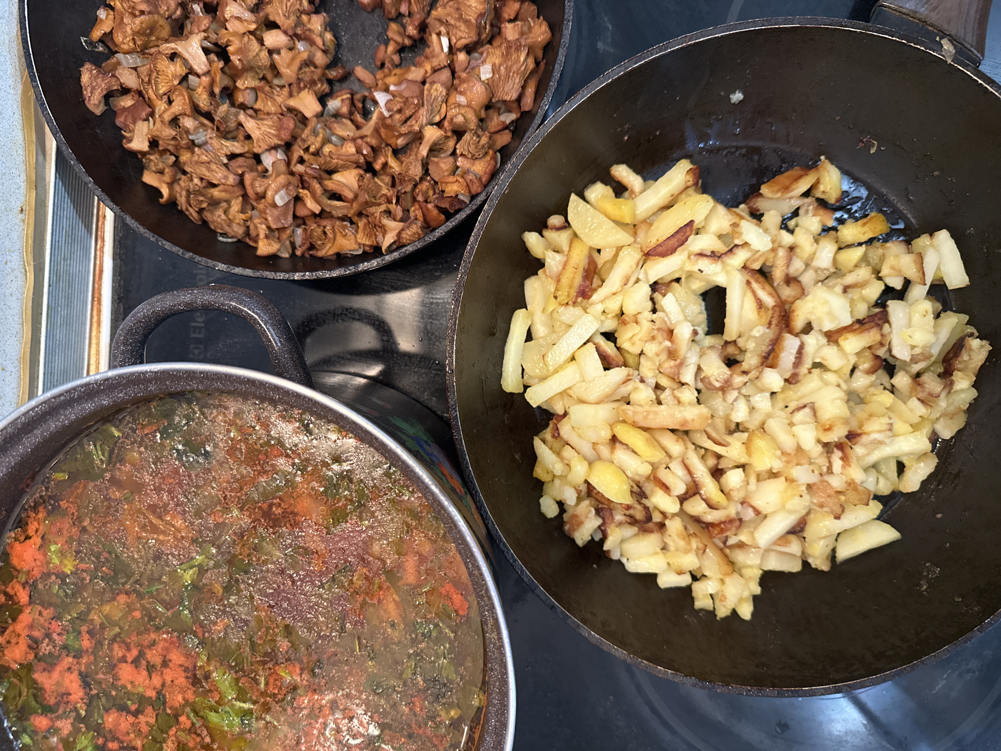A close-up, overhead shot shows three pans on a black stovetop. In a large pot on the left, a reddish-orange soup with green herbs is simmering. To the right of the soup pot, a frying pan is filled with sautéed mushrooms and diced onions. Below these, another frying pan contains golden-brown fried potatoes, cut into strips and cubes. The overall scene suggests a home-cooked meal being prepared. The stovetop has markings suggesting it is a brand like Electrolux. There is no clear indication of the location within Sverdlovskiy, Russia.