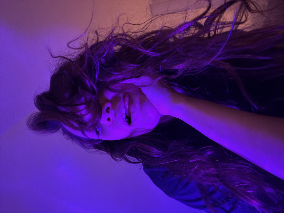 An angled close-up captures a person with long, flowing dark hair, illuminated by a vibrant blue light. The individual's face is partially obscured by their hair. They are sticking out their tongue with their head tilted slightly. Their hand is touching their cheek. The overall tone is both playful and mysterious, enhanced by the strong color cast. The background is a light color.