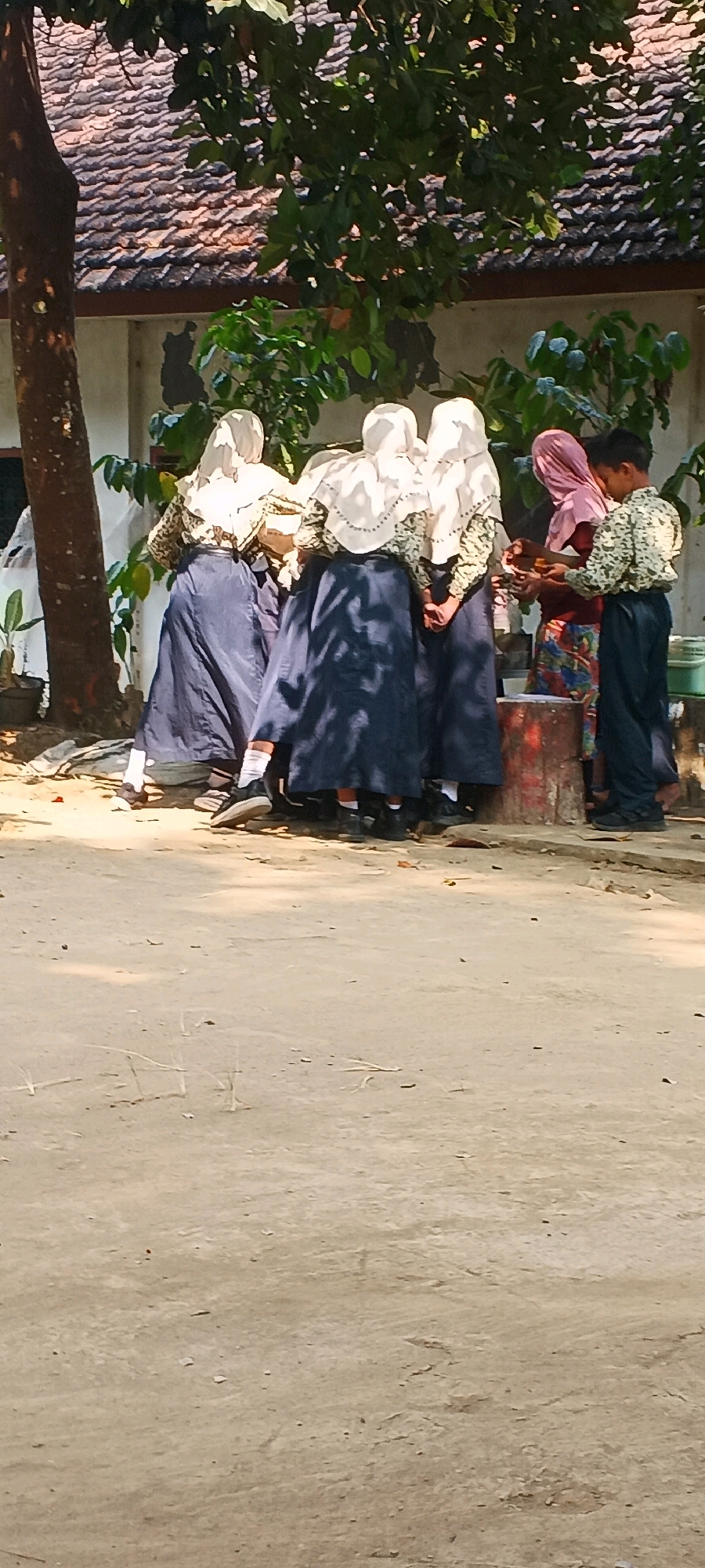In Penanggungan, Indonesia, a group of five students, three girls and two boys, are gathered around a reddish-brown cylindrical object. The girls are wearing white hijabs and dark blue skirts, with one in a patterned top and the others in white. The boy on the right is wearing a light-colored patterned shirt and dark pants. The other boy is partially visible on the far left. They appear to be interacting with something small held in their hands. The setting is outdoors, with a building and a tiled roof in the background, partially obscured by trees with green foliage. The ground is sandy and uneven, with some scattered leaves. The scene suggests a school environment or a communal gathering.