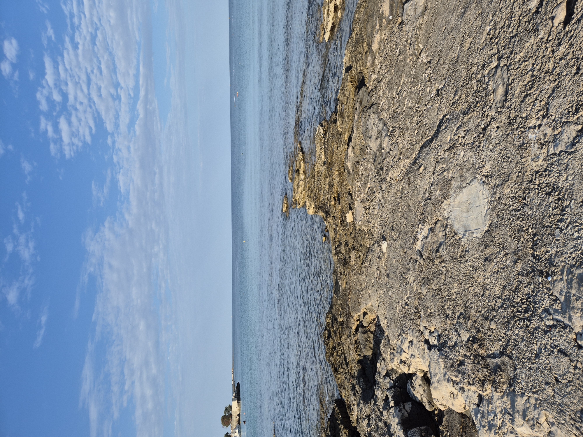The image shows a rocky coastline in Zambratija, Croatia, with the Adriatic Sea stretching out to the horizon under a partially cloudy blue sky. In the foreground, rough, textured rocks and sand dominate the scene. A small inlet of calm, blue water is visible to the right. In the distance, a line of buildings and vegetation can be seen along the shore to the left. Several buoys and possibly a distant sailboat are visible on the sea. No people are clearly discernible in the image. The light suggests it is daytime, likely late afternoon given the soft shadows.