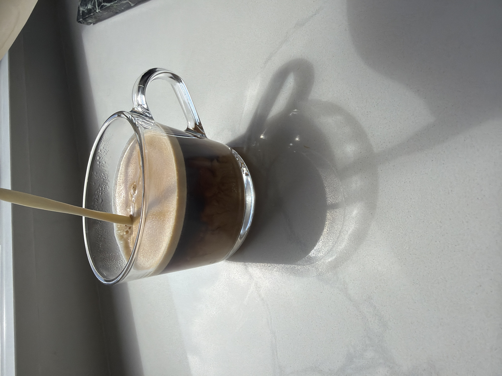 This is a high-angle, close-up, rotated shot of a clear glass mug filled with coffee and a creamy layer of frothed milk being poured into it. The mug is resting on a light-colored, speckled countertop, possibly marble or granite. The background shows a gray wall and a shadowy area, suggesting indoor lighting. The shadow of the mug and its handle is cast on the countertop, indicating a light source from the side and slightly above. The coffee itself has a dark brown base with a lighter brown, frothy layer on top, indicating a latte or a similar coffee beverage is being prepared. A slender straw or stick is submerged in the coffee, pouring the milk into the mug. The image conveys a sense of calm and domesticity, possibly capturing a morning routine or a moment of relaxation. There is no visible text in the image. The scene is static, with the focus on the beverage preparation. The lighting suggests it could be daytime, with natural light contributing to the shadows. The clean aesthetic of the countertop and the modern design of the mug and straw hint at a contemporary kitchen or coffee nook setting.