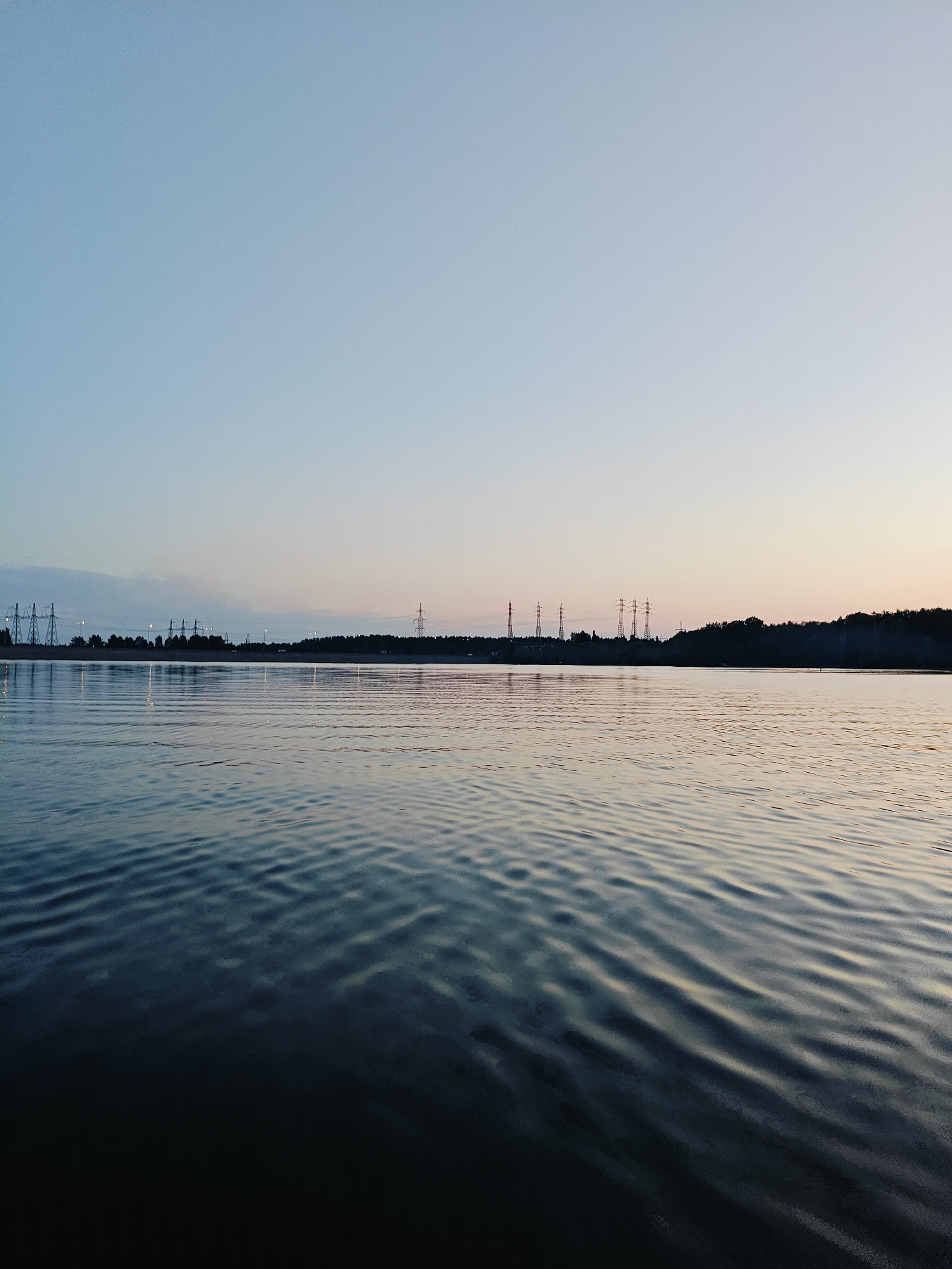 This is an outdoor, low-angle, full shot image of a calm body of water at dusk, likely in Noviy, Russia. The water's surface is disturbed by subtle ripples, reflecting the pale sky and the dark silhouette of a tree line in the distance. Several tall, lattice-like power transmission towers are visible against the horizon, punctuating the natural landscape with industrial infrastructure. The sky is a soft gradient of blue to pale yellow near the horizon, indicating the end of daylight. There are no people or animals visible in the scene.