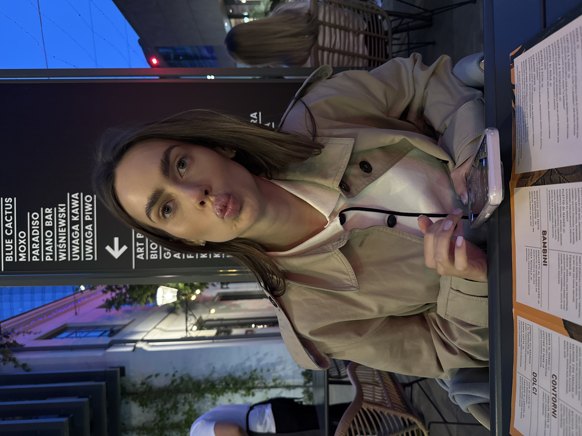 A woman is sitting at a table outside a restaurant or bar. She is wearing a beige trench coat over a white top with black trim and is holding a phone. Her lips are puckered as if she is about to kiss. On the table are menus with Italian text. The background shows a street scene with buildings lit by streetlights and neon signs. The signs indicate the names of bars and businesses, including "BLUE CACTUS", "MOXO", "PARADISO", "PIANO BAR", "WIŚNIEWSKI", "UWAGA KAWA", "UWAGA PIWO", and "ART P". There is also an arrow pointing towards "BIO" and "G...". The exact location in the city of Unknown, Unknown, cannot be determined with certainty from this image.