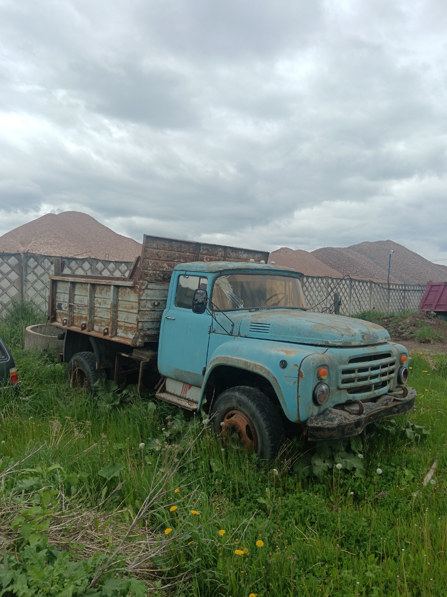 An old, blue, rusted truck sits in a field of green grass and dandelions on an overcast day in Яльгелево, Russia. The truck has a rusty metal bed and appears to be non-functional, with overgrown vegetation surrounding it. Behind the truck, a concrete fence borders a large pile of sand or gravel. Another vehicle, possibly a car, is partially visible on the left. The sky is filled with grey clouds.