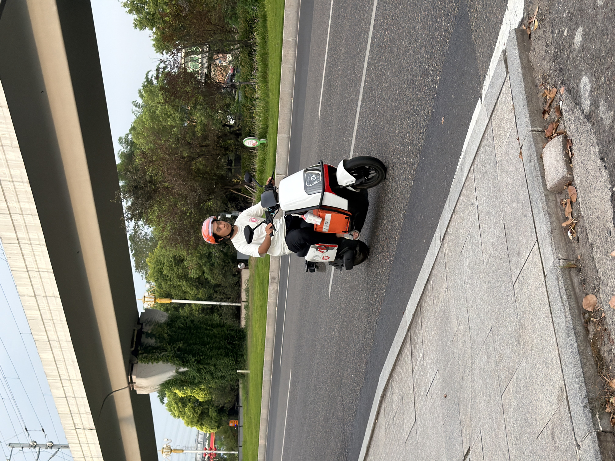 A man wearing an orange helmet and a white t-shirt is riding a white scooter on a paved road in Jiangjin, China. The scooter has an orange bag attached to the back. In the background, there is a large elevated concrete structure with power lines, trees, and streetlights. The foreground shows a gray sidewalk and a road with white lane markings. The sky is overcast.