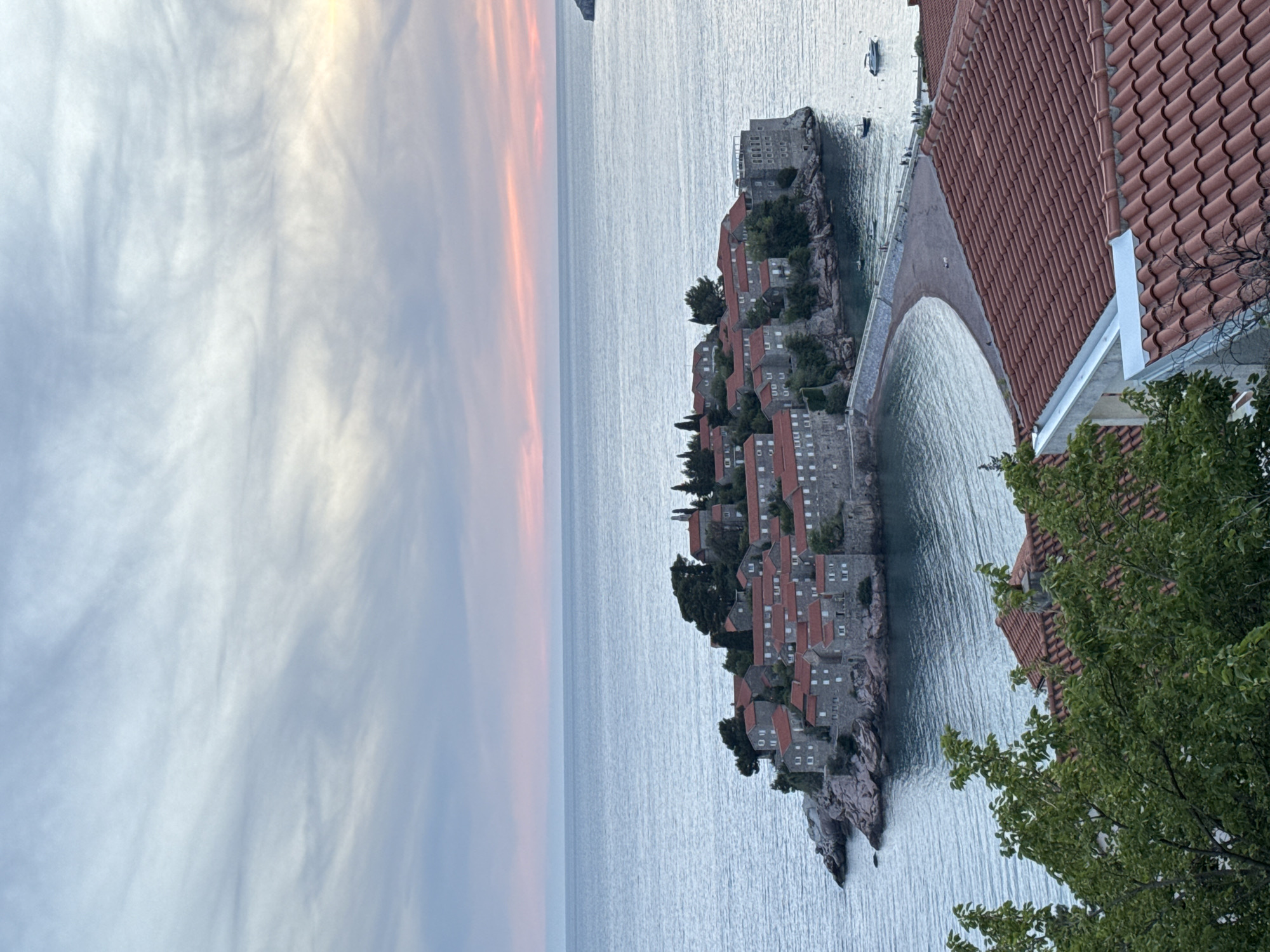 The image depicts Sveti Stefan, Montenegro, a small islet connected to the mainland by a narrow isthmus. The islet is densely packed with buildings having red-tiled roofs, typical of the Adriatic coast architecture. These buildings are constructed on rocky terrain that forms the base of the islet. The isthmus appears to be a concrete causeway allowing access from the mainland. The surrounding water is calm and appears to be part of the Adriatic Sea.
On the right side of the frame, part of a building with a similar red-tiled roof and white trim is visible. Green foliage from trees or bushes is also visible in the foreground, partially obscuring the view. The sky shows a gradient of colors, transitioning from blues and grays to pink and orange hues, suggesting either sunrise or sunset.
