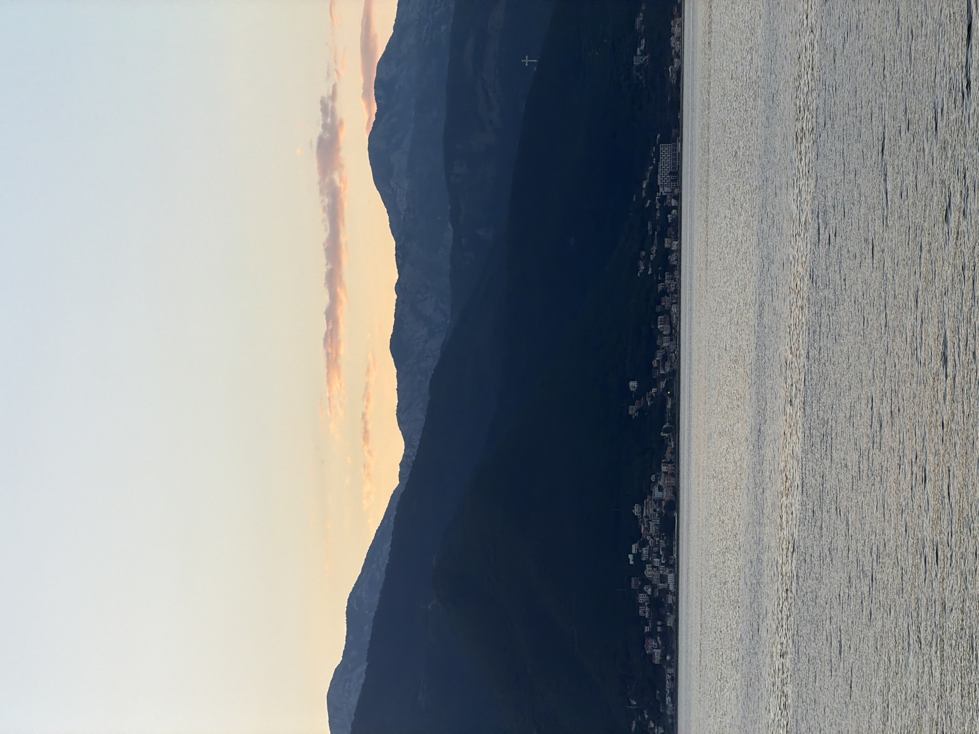 The image depicts the coastline of Gošići, Montenegro, at either sunrise or sunset. The sun, positioned on the left, illuminates the sky with pale orange hues. A mountain range runs down the center of the frame, partially obscuring the sky. The city of Gošići is nestled along the base of the mountains, on the shoreline. A body of water, likely the Adriatic Sea, occupies the right half of the image, its surface reflecting light. A prominent white cross is visible on the highest point of the mountain range.