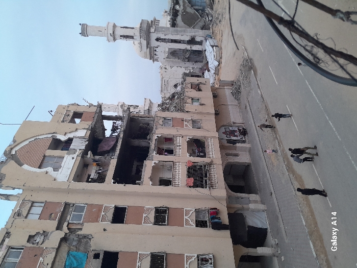 This image depicts a scene of extensive structural damage in Khan Yunis, Palestinian Territory. The foreground is observed from an elevated perspective, showcasing a paved road with white lane markings. Approximately eight individuals, primarily adult males and at least one child, are present on the street and adjacent sidewalk, appearing to be walking or standing in small groups.
To the left, a multi-story building, at least six floors high, exhibits severe destruction. Large sections of its beige facade are missing, exposing internal rooms, broken windows, and twisted rebar. Balconies with dark brown railings are visible, some with clothes hanging to dry. An arched decorative element on an upper floor is partially intact. Debris is scattered around its base. On the opposite side of the street, to the right and further back, stands a white mosque with a tall minaret and a dome, both bearing visible damage including holes and missing sections. Surrounding the mosque are piles of rubble and what appear to be makeshift shelters constructed from tarps. The sky above is overcast or hazy.