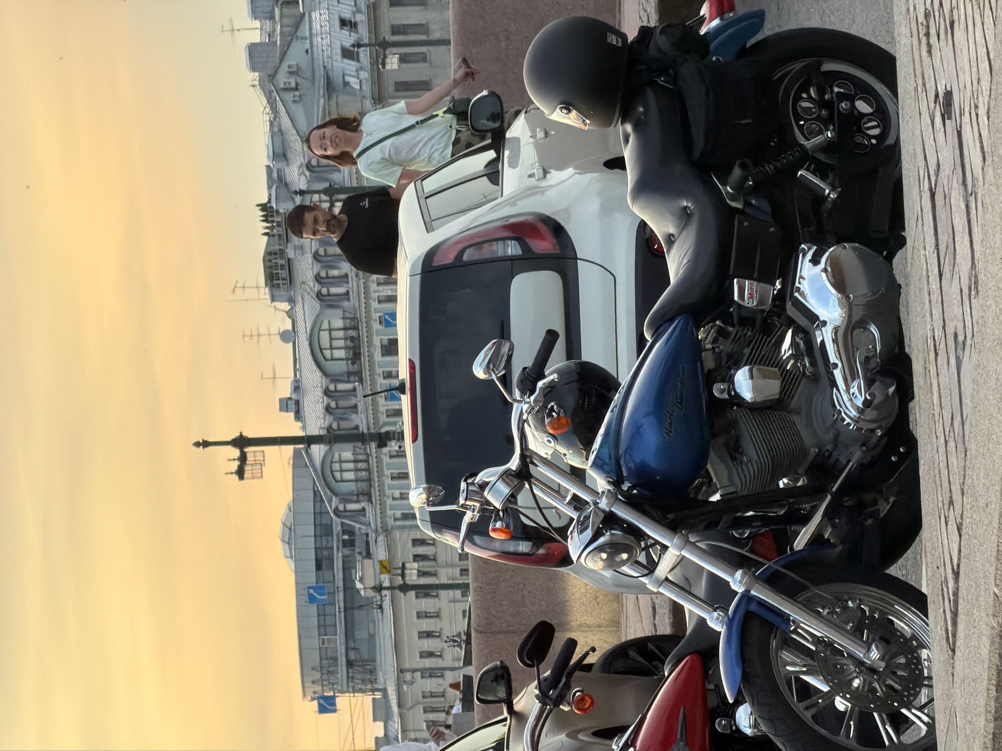 This is a medium shot of several parked motorcycles and two people standing behind a white car, with buildings and the sky in the background, likely in Saint Petersburg, Russia. The sky is a warm yellow, suggesting it is either sunrise or sunset.
In the foreground, a blue Harley-Davidson motorcycle is prominently displayed, with its front wheel, engine, and handlebars visible. A portion of a red motorcycle is visible to the left. A black helmet rests on the seat of the blue motorcycle.
Behind the motorcycles and to the right is a white car. Standing next to and slightly behind the car are a man and a woman. The man is wearing a black t-shirt, and the woman is wearing a light-colored t-shirt and camouflage pants, with a green bag slung over her shoulder. They appear to be looking towards the viewer or slightly to the right.
In the background, various buildings with distinctive architecture can be seen, including a building with a curved roof and large windows. A lamppost with two lights is also visible. The overall setting appears to be an urban environment, possibly a waterfront area, given the presence of a stone embankment in the foreground. The scene captures a moment of quietude with the parked motorcycles and the two individuals observing the surroundings.