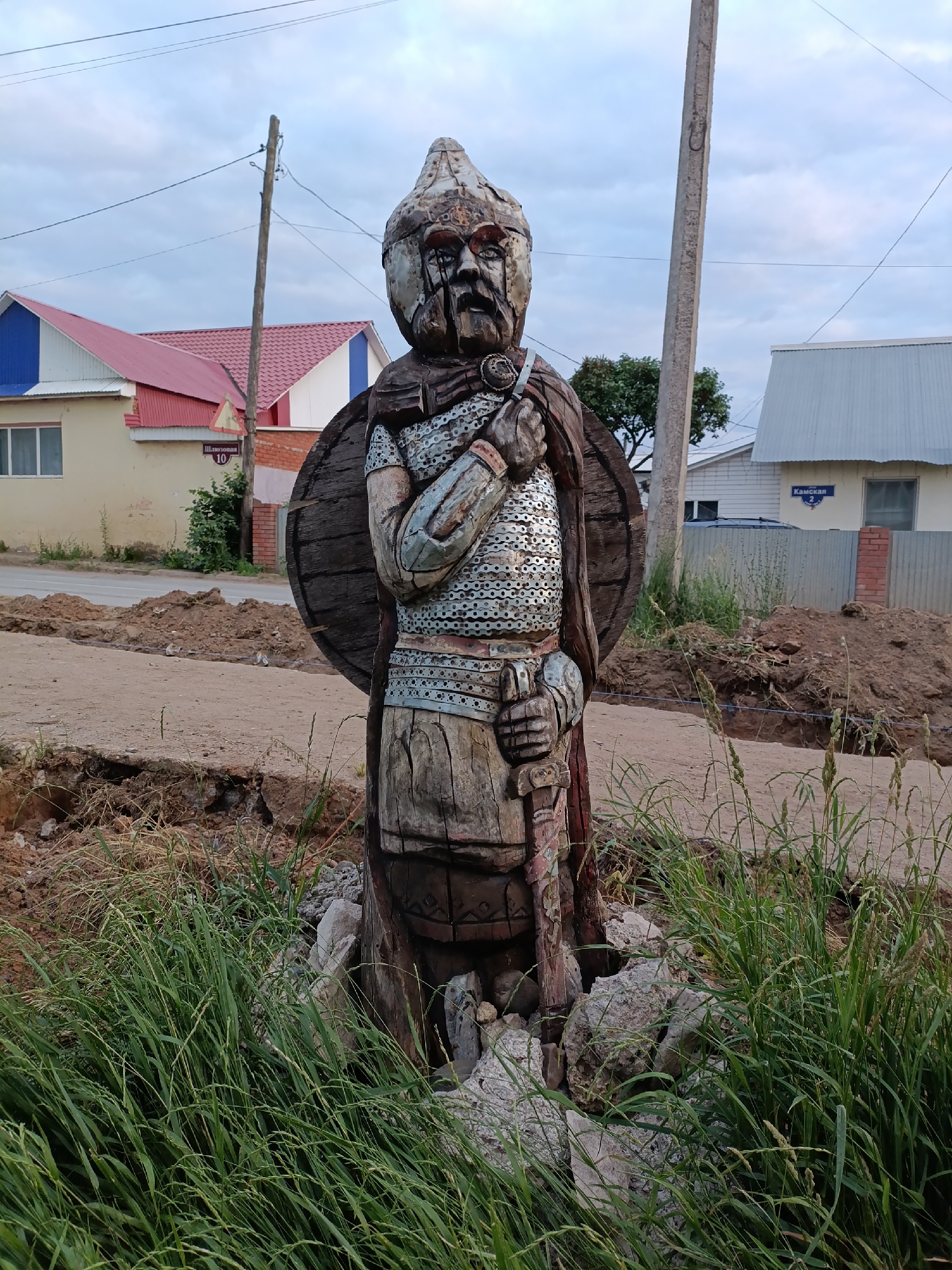 A wooden sculpture of a medieval warrior stands on a roadside in Chaikovsky, Russia. The warrior is depicted wearing a helmet, chainmail armor, and a cape, and is holding a sword and a shield. The sculpture is surrounded by grass and rocks, and in the background, there are houses and a cloudy sky. The street is unpaved and appears to be under construction, with piles of dirt and debris visible. A street sign in the background indicates "Шлюзовая 10" (Shlyuzovaya 10), which is likely the street name and house number. Another street sign visible says "Камская 2" (Kamskaya 2).