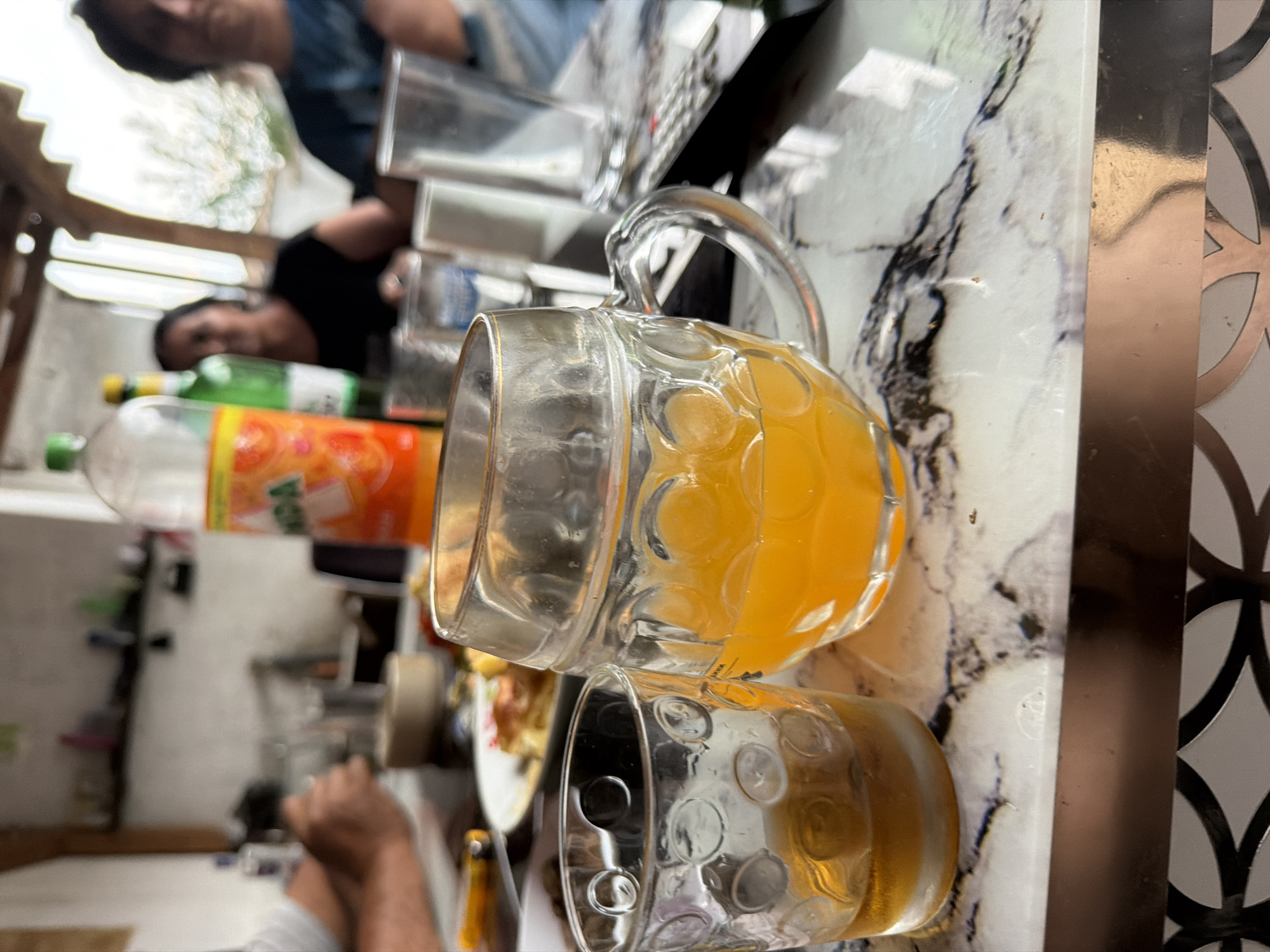 The image displays an indoor or semi-outdoor dining scene in Dabene, Bulgaria. A white and black marble-patterned tabletop occupies the foreground. On this surface, a small, clear glass tumbler with a textured circular pattern contains an amber-colored liquid on the left. To its right, a larger, clear glass mug with a handle and similar textured pattern holds a bright orange liquid. Below the tabletop, a reflective metallic trim with a dark, patterned border is partially visible.
In the midground, behind the beverages, a blurred white plate with a baked or pastry-like food item is visible, alongside a yellow-handled utensil. Additional blurred glassware and table accessories are present.
The background is out of focus, revealing several individuals and environmental details. To the far left, a person wearing a white short-sleeved top has their arm and hand visible near the table. A white wall with a dark shelf holding indistinct objects is positioned behind them. In the center, a clear plastic bottle with an orange and yellow label displaying the text "ANDA" (likely Fanta) stands upright, adjacent to a partially visible green glass bottle with a yellow cap. Two individuals are seated towards the right background; one person with dark hair wears a black top, and another person with dark hair wears a blue patterned top. Both faces are blurred. Behind these individuals, a brightly lit and heavily blurred outdoor scene, possibly featuring foliage, suggests an open window or archway. Blurred wooden structural beams are visible overhead. The overall scene suggests a casual social gathering or meal.