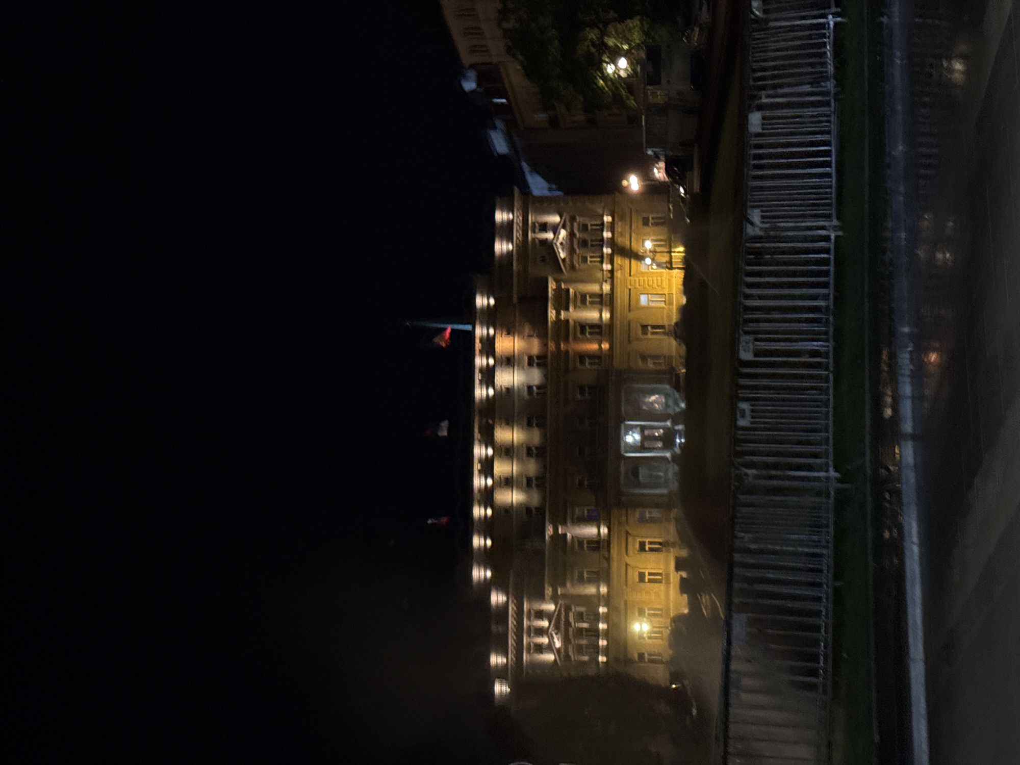 At night, a lit building is reflected in water. The building is located in Belgrade, Serbia. The area around the building is dark. In the foreground is a bridge.
