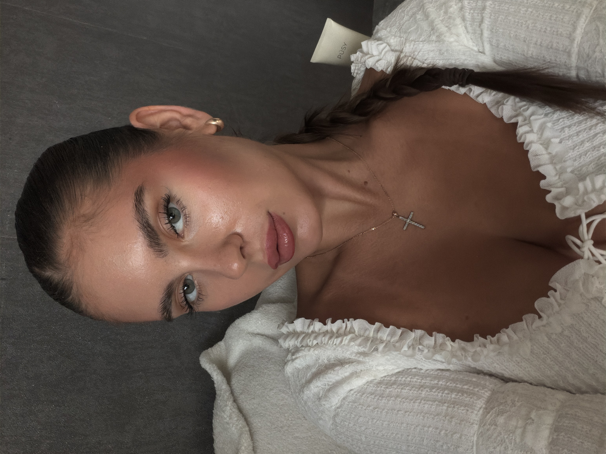 A woman with light eyes and brown hair is photographed close-up, partially lying down. She is wearing a white textured garment with ruffled edges and a delicate gold necklace with a cross pendant. Her hair is styled in a braid. In the background, partially visible, is a white tube labeled "PUSY". The surface she is lying on appears to be a dark grey, possibly concrete or tile. The location and specific activities are not visible in the image. The location in the city of Krasnoyarsk, Russia is not discernible from the image.