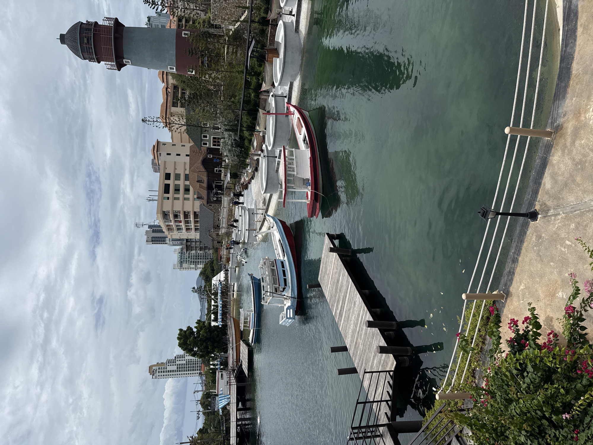 This image depicts a canal-like waterway in Pattaya City, Thailand, with several boats moored. On the right side of the image, a tall, lighthouse-like structure with red and grey sections dominates the background. Several multi-story buildings, typical of a city environment, are visible behind the lighthouse and along the canal. The waterway is lined with pathways and low walls, and a wooden dock extends into the water on the left. Various small fishing boats, painted in white, red, and blue, are floating in the water. Some of these boats are decorative, while others appear functional. There are a few people visible in the distance walking along the pathways. The water is a murky green, reflecting the overcast sky. Lush green vegetation and pink flowers are present in the foreground on the left, adding a touch of nature to the urban setting.