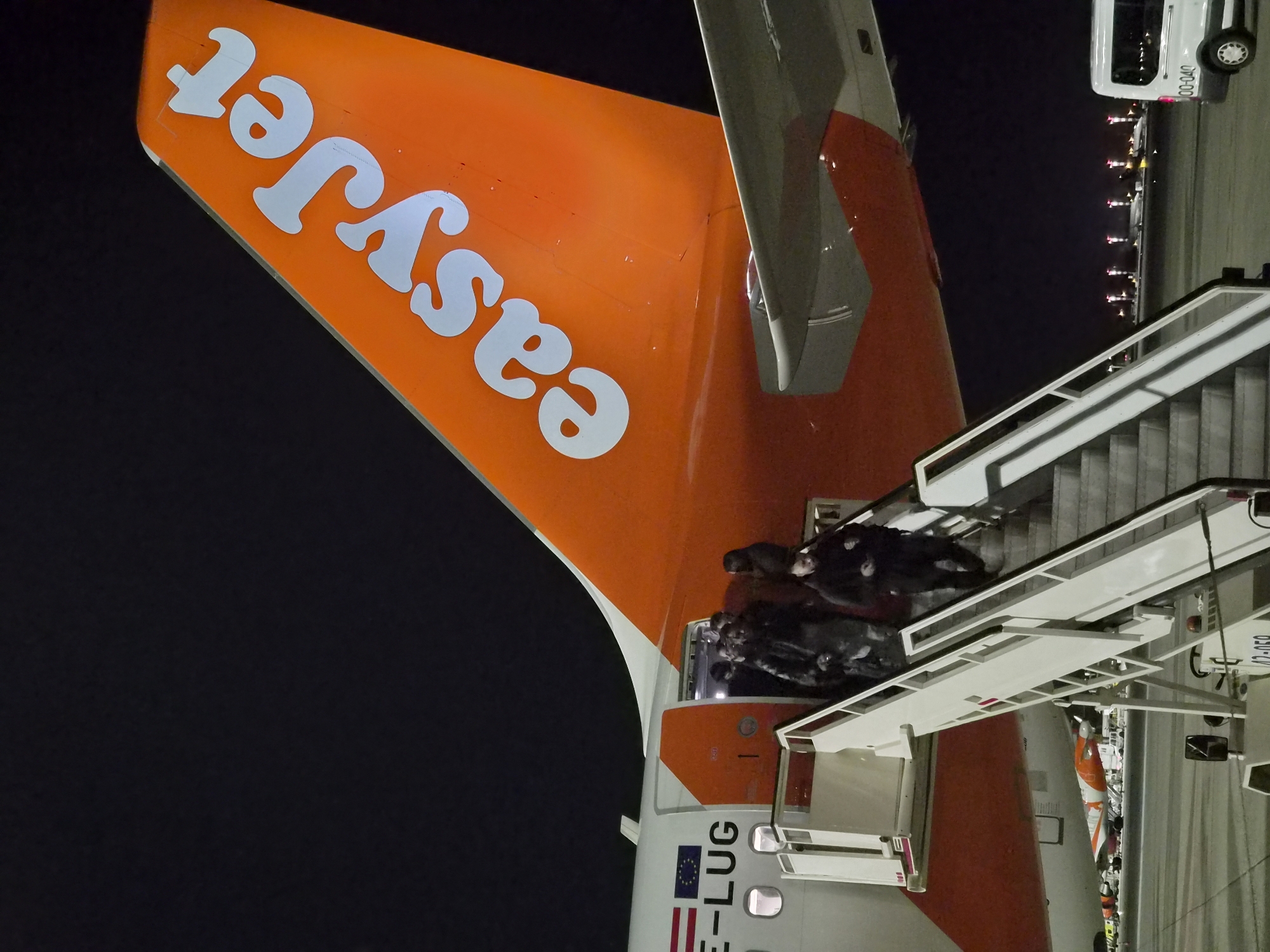 At night, an EasyJet airplane is stationed at Vila Nova da Telha, Portugal. Passengers are exiting the aircraft via mobile stairs leading to the ground. The EasyJet logo is prominently displayed on the orange tail of the plane. A white van is visible in the background, parked near runway lights.