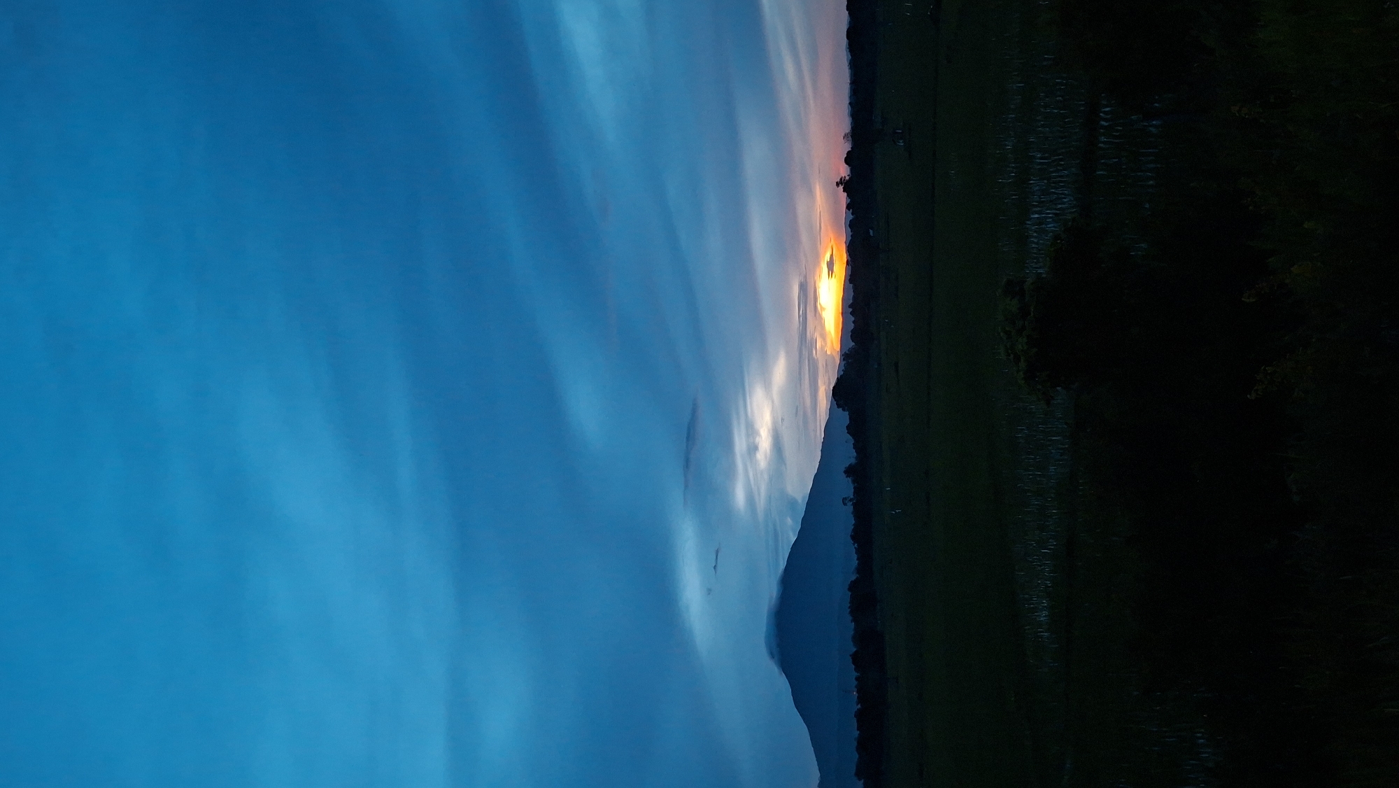 The image depicts a tranquil sunset scene over a rural landscape in Baksukon, Indonesia. The sky is a deep, vibrant blue with streaks of orange and yellow where the sun is setting behind a distant mountain range. The mountain, silhouetted against the colorful sky, is partially obscured by clouds. In the foreground, a dark, vast expanse of rice paddies stretches across the land, reflecting the dimming light. The overall impression is one of peacefulness and natural beauty, characteristic of the Indonesian countryside at dusk. There are no people or man-made structures clearly visible, emphasizing the natural and serene atmosphere of the location.