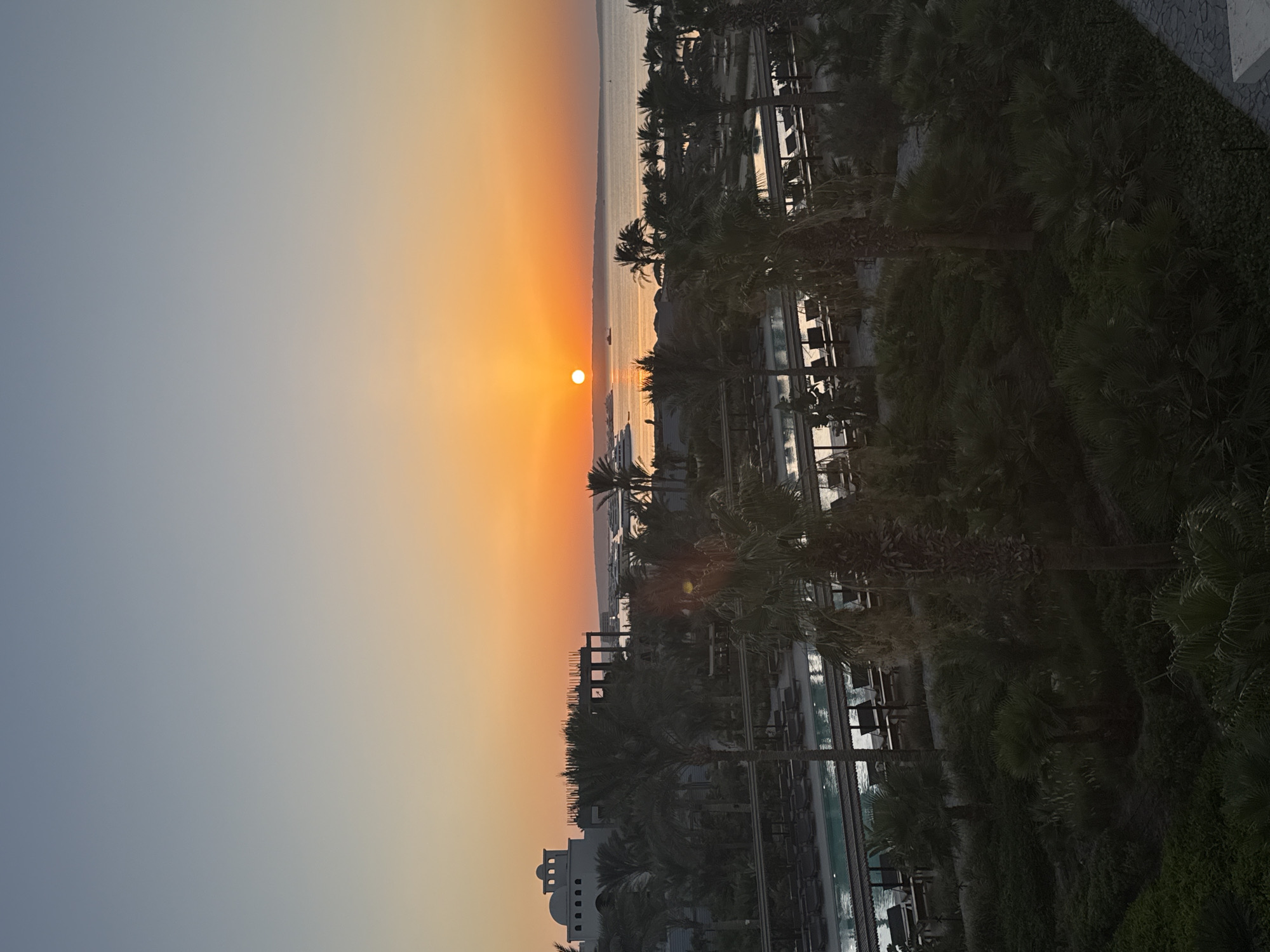 The image depicts a serene sunset over the city of Hurghada, Egypt. The sky is a gradient of warm oranges and yellows near the horizon, transitioning into a soft blue at the top. The sun, a bright orange orb, is low in the sky, casting a gentle glow over the scene.
In the foreground, lush green palm trees and dense foliage create a tropical ambiance. Beyond the vegetation, a swimming pool area is visible, with rows of empty lounge chairs arranged under shaded structures, suggesting a resort or hotel setting. The water in the pool reflects the warm hues of the sunset.
Further in the distance, several buildings can be seen, including a distinct white structure with arches, possibly a hotel or administrative building, and another structure appearing to be under construction. The calm sea stretches across the horizon, dotted with a few boats and a large yacht. The overall atmosphere is peaceful and tranquil, characteristic of a coastal resort town during sunset.