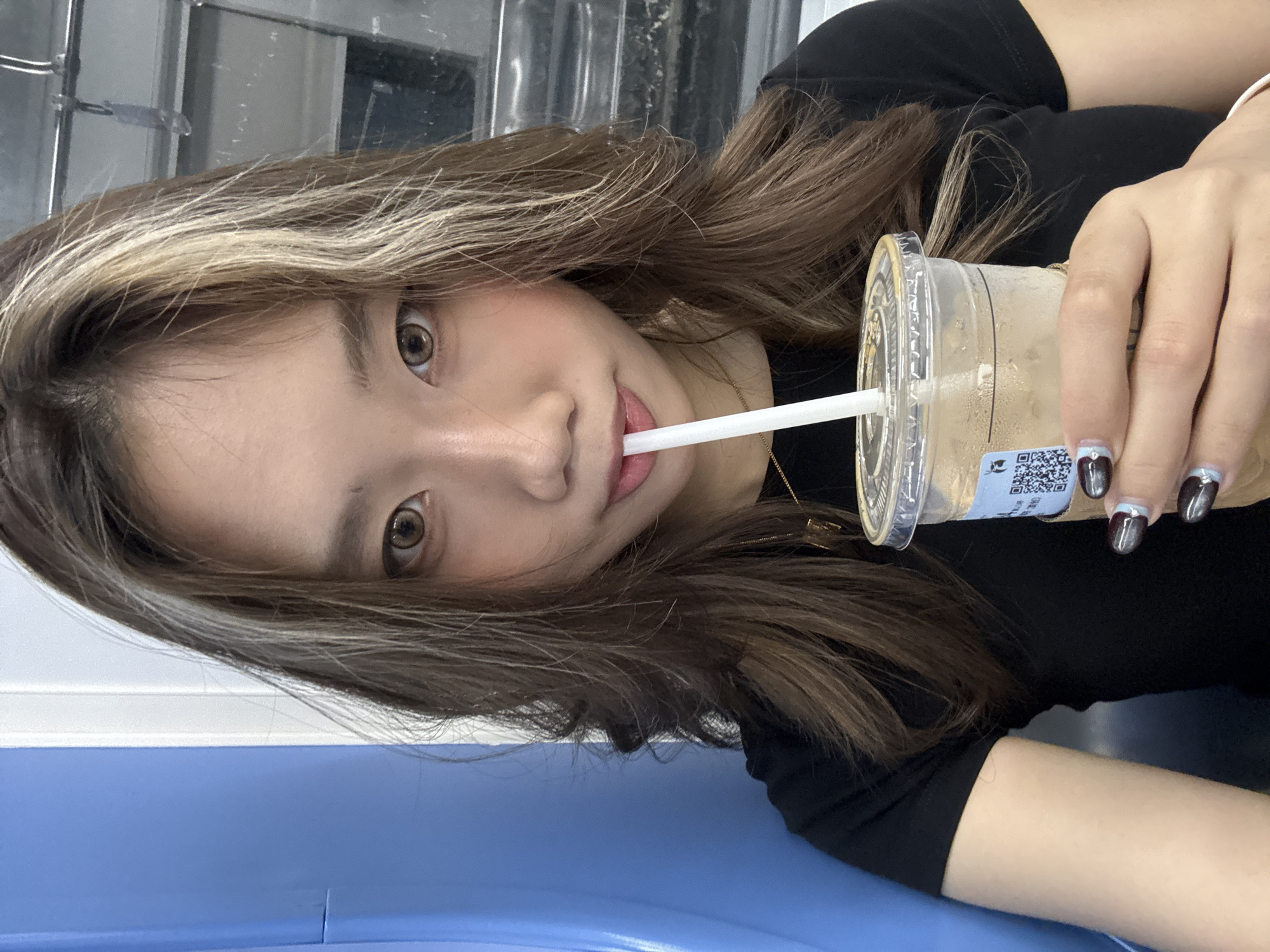 A person with light brown hair with blonde highlights is holding a plastic cup with a straw, possibly a beverage, while sitting on a blue surface. They are wearing a black top and have manicured nails. The location is Nan'an District, China. A metal structure is visible in the background.