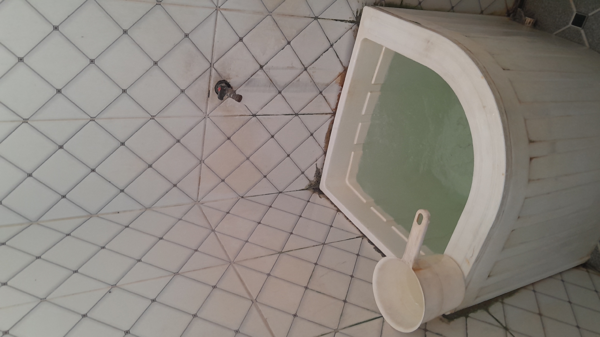 In Sambong, Indonesia, a bathroom scene is depicted, focusing on a corner bathtub filled with light green water. A white plastic ladle rests on the edge of the tub, partially submerged in the water. The walls are covered in white square tiles with a diamond pattern, and a silver faucet is visible on the tiled wall above the tub. The floor has grey and white tiles arranged in a diamond pattern as well. The scene appears to be indoors, likely in a residential setting.