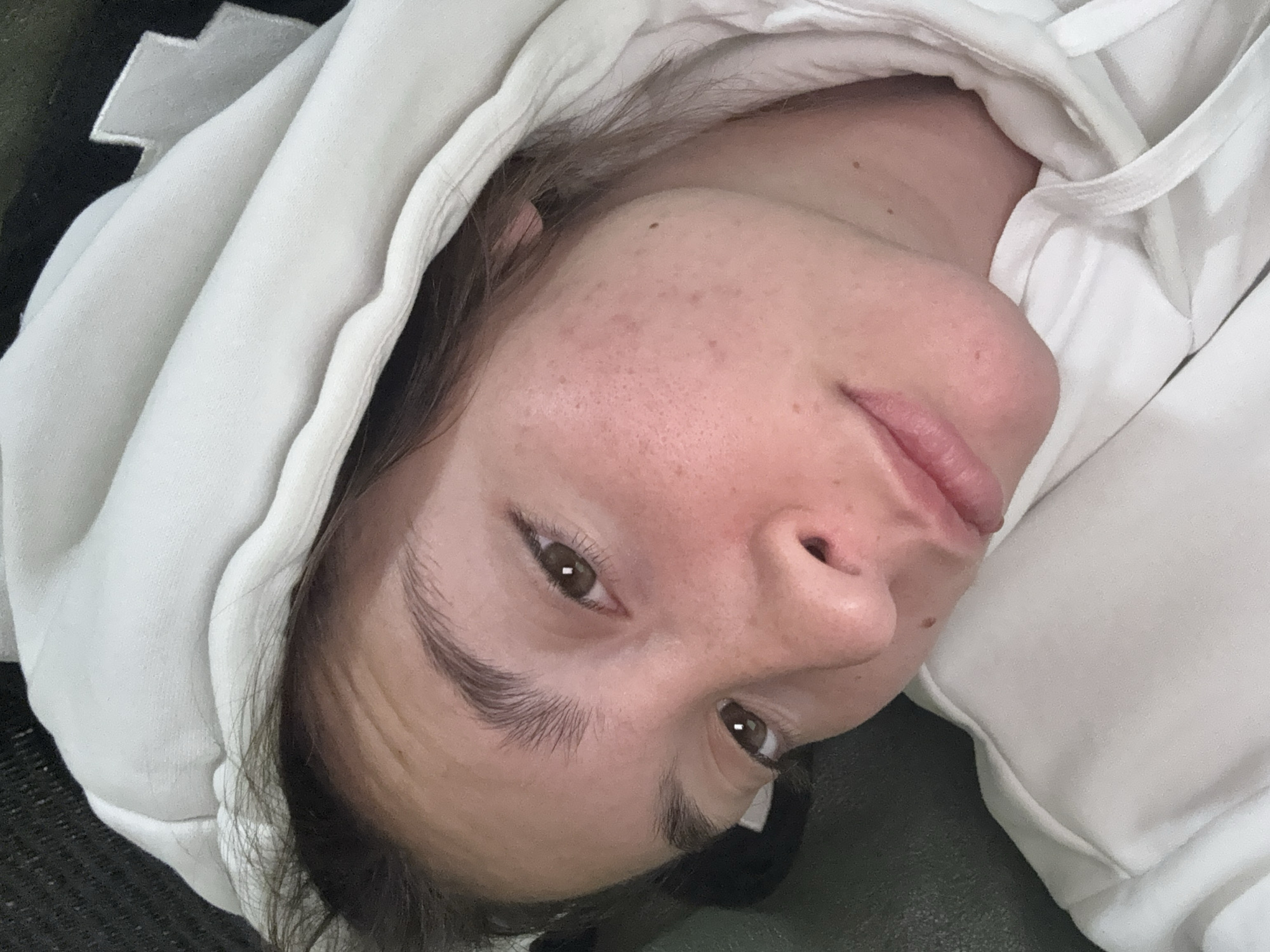 A close-up, overhead shot captures a young person wearing a white hooded sweatshirt, their face tilted to the side. The person has brown eyes and fair skin with some freckles and redness. Their hair is dark brown. The background is dark and out of focus, with a hint of a textured surface on the left. The setting appears to be indoors, possibly a living room or bedroom, and no specific location in Sofia, Bulgaria can be determined from the visual information provided.