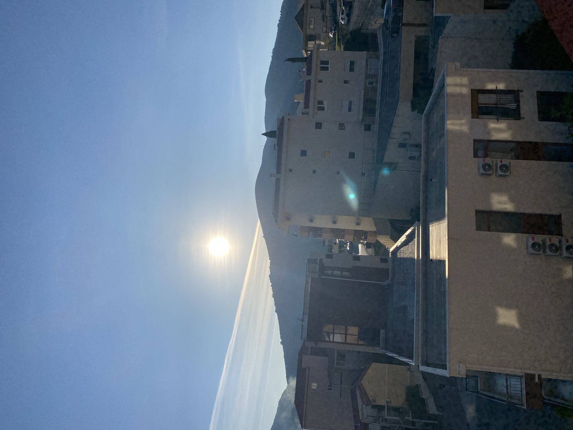 The image depicts a sunny scene in what appears to be Budva, Montenegro, given the architecture and mountainous backdrop. Tall apartment buildings are visible, suggesting a relatively dense urban environment. The sun is prominent in the sky, casting a glare and creating strong shadows on the buildings. The mountains rise in the background, partially obscured by haze. The sky is clear blue. There are no people visible in the image.