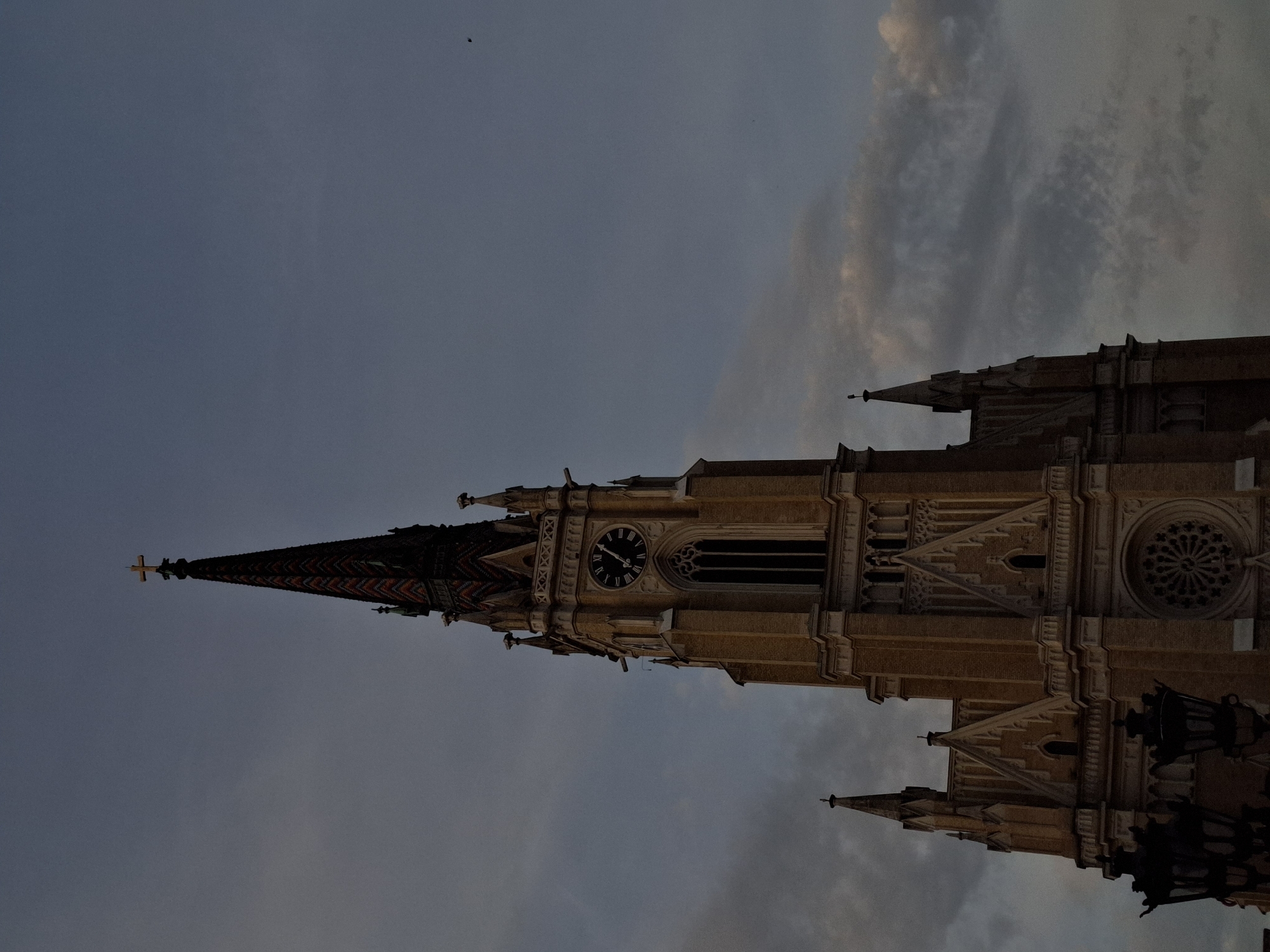 The image captures a dramatic, low-angle view of the towering Gothic Revival spire and upper facade of the Name of Mary Church (also known as the Cathedral of Novi Sad) in Novi Sad, Serbia. The church dominates the frame, stretching towards a twilight sky.
The main spire, topped with a golden cross, features a distinct patterned roof composed of colorful tiles. Below the spire, a large, circular clock face with black Roman numerals (I through XII) and hands is prominently displayed, indicating approximately 7:00 or 7:05. Adjacent to the clock tower, the church exhibits intricate architectural details, including tall, arched windows, decorative pinnacles, and a large rose window with ornate tracery on a section to the right. The building's facade is constructed from light-toned stone or brick, characteristic of its architectural style.
The sky is a muted, deep blue-grey, with softer, lighter clouds visible on the right, suggesting the scene is set during dusk or dawn. The lighting is subdued, contributing to a solemn and grand atmosphere. A small, dark, indistinct object, likely a bird or insect, is visible against the sky in the upper left. No people or other textual elements beyond the clock numerals are visible in the photograph, keeping the focus solely on the magnificent architecture and the atmospheric sky.