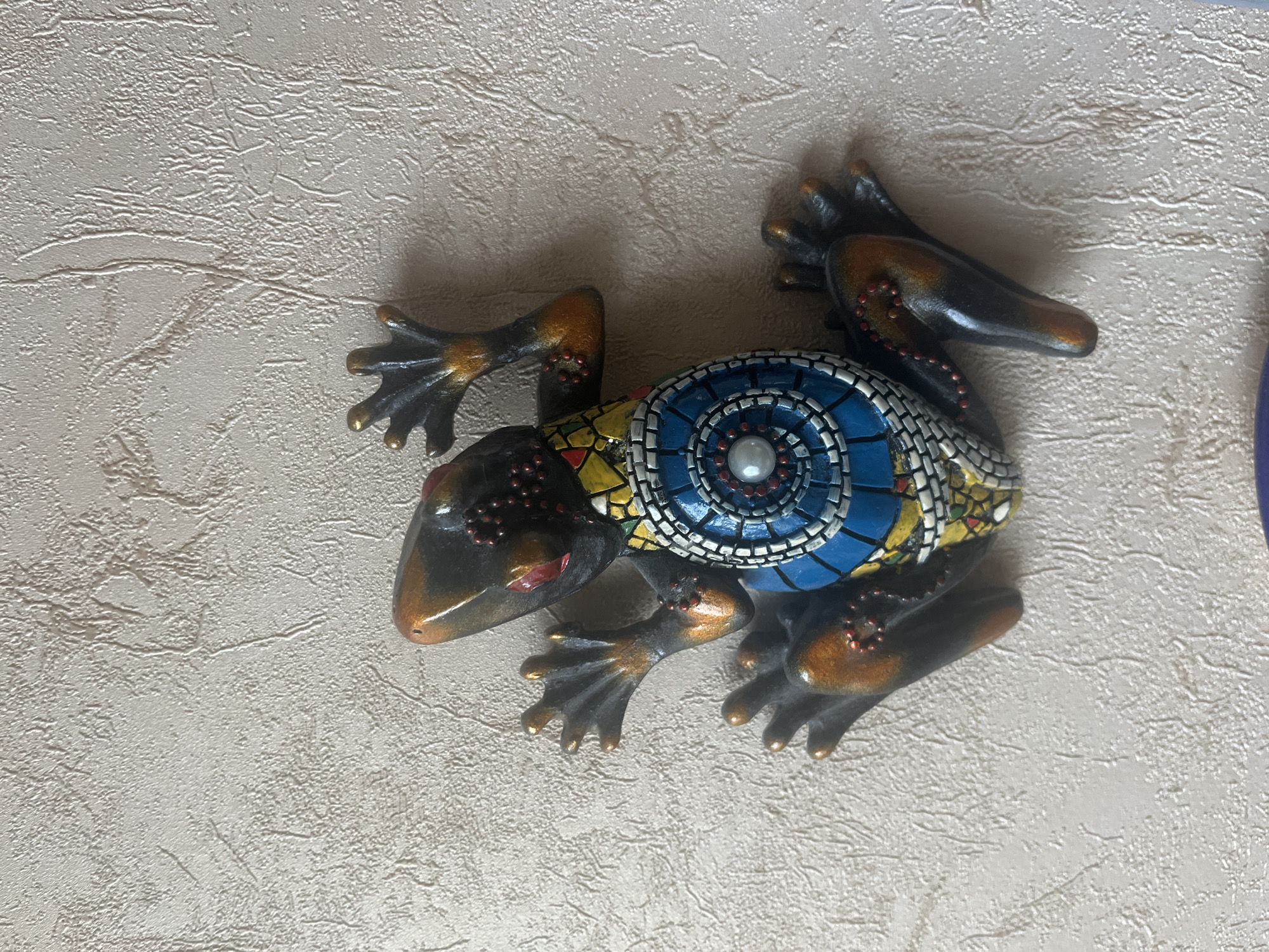 A decorative ceramic lizard is affixed to a textured beige wall. The lizard is dark with orange and metallic accents. Its back features a circular mosaic design with blue, yellow, and white tiles, and a pearl-like embellishment in the center. Small red beads dot the lizard's body. The lizard's limbs are splayed outwards, appearing to be clinging to the wall. There is no indication of a specific location within Vladimirovka, Russia.