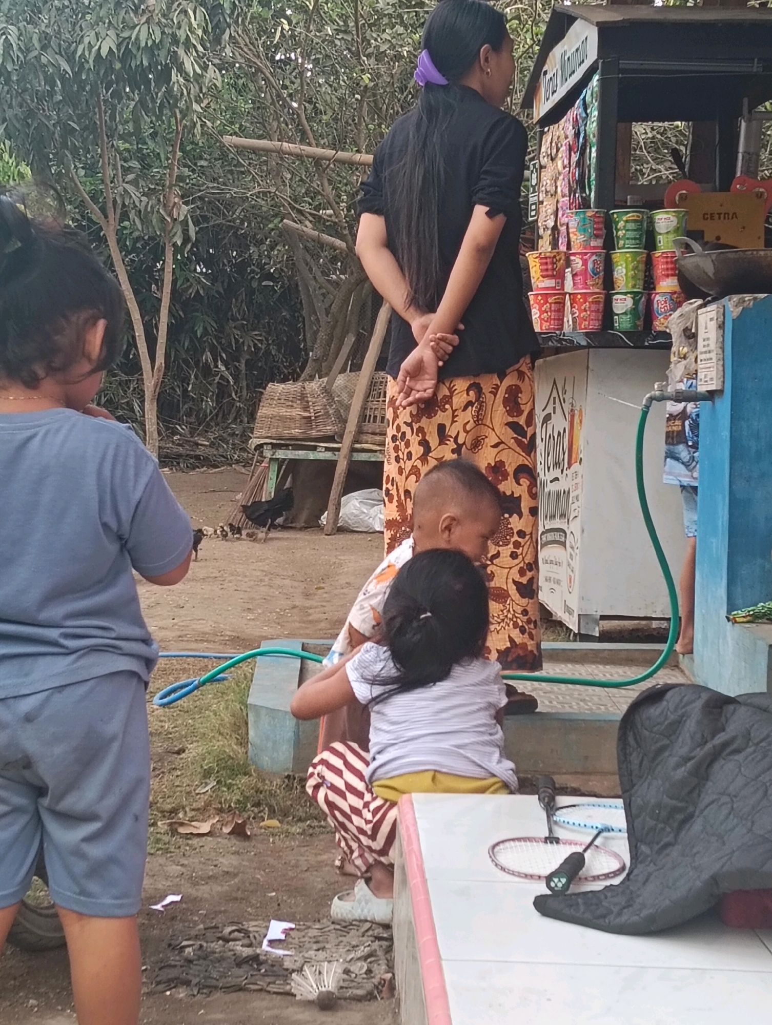 In Kasiyan Timur, Indonesia, a scene unfolds near a small outdoor vendor stall. A woman stands with her back to the viewer, her long dark hair pulled back with a purple accessory, wearing a black shirt and an orange and brown patterned skirt. In front of the stall, which displays various colorful packaged goods and some food items, a young child is being held by another person, who is mostly obscured from view. To the left, another child in a blue t-shirt and shorts stands facing away from the camera. A green hose is connected to a faucet on a blue structure. In the background, there are trees and a woven basket on a wooden structure, and a few chickens are present on the dirt ground. The overall setting appears to be a casual outdoor environment, possibly a residential area or a roadside market.