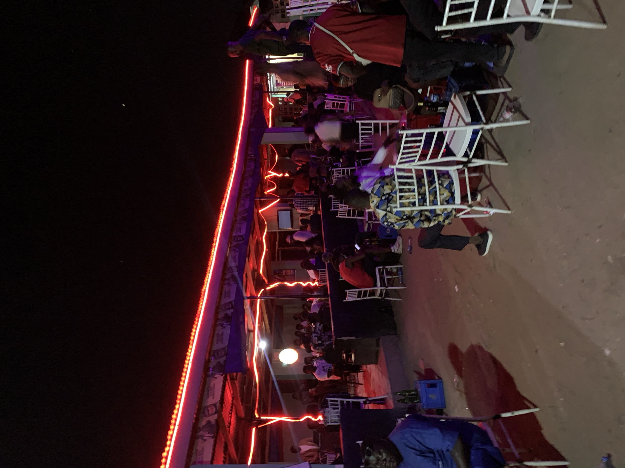 The image shows a crowded outdoor bar or restaurant in Kabong, Nigeria at night. The structure has a roof outlined with red neon lights. People are seated at tables and chairs, mostly white plastic. There are drinks and bottles visible on some of the tables. The area outside the seating is dirt or sand. The sky is dark.