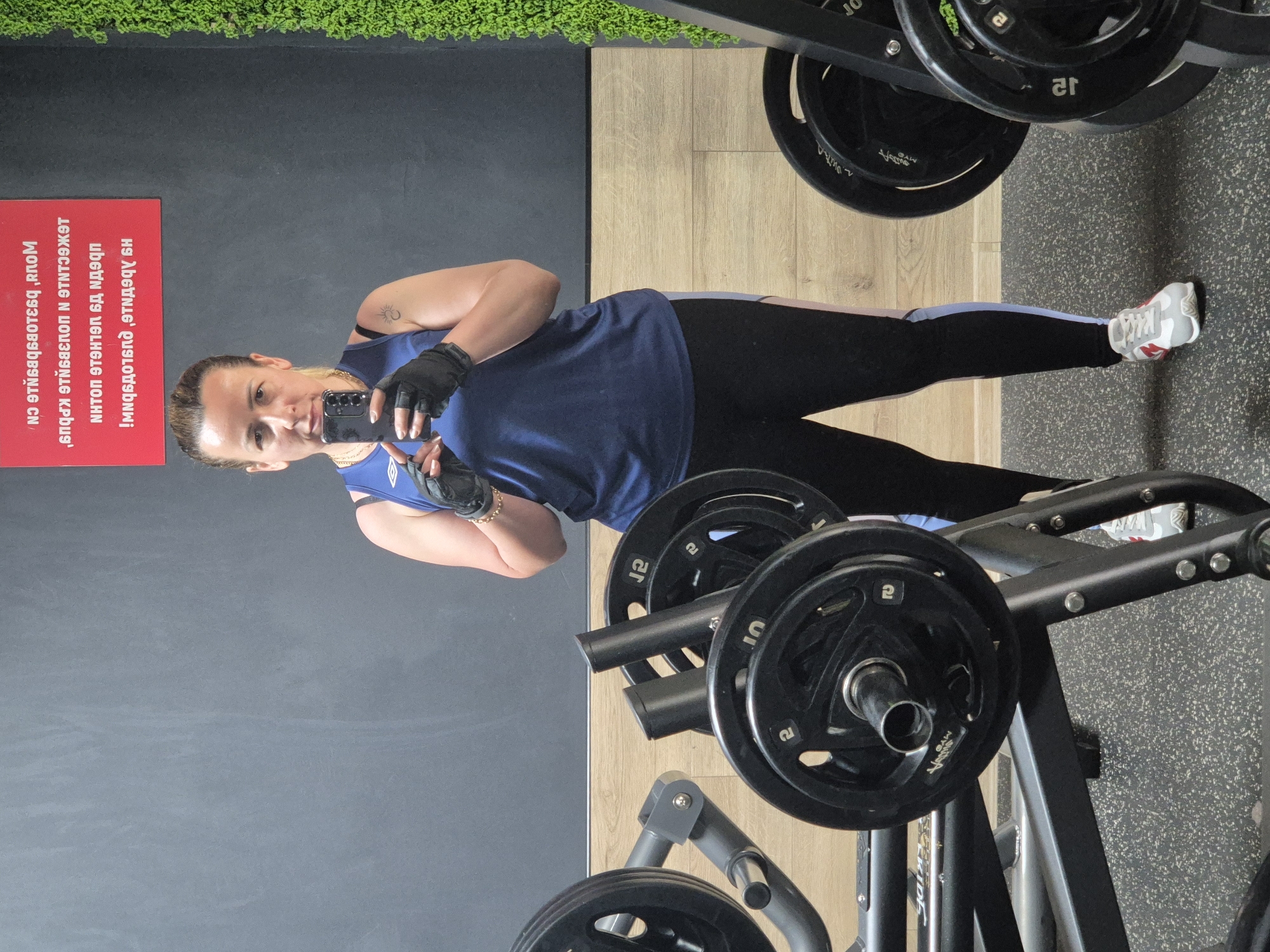 A woman stands in a gym, taking a mirror selfie. She's wearing a dark blue tank top, black workout leggings with a light blue stripe, black gloves, and white sneakers with red accents. Her hair is pulled back, and she has a tattoo of a sun and moon on her upper arm. Behind her, weight plates are stacked on a rack. A red sign with white text hangs on the wall, its reflection visible in the mirror.