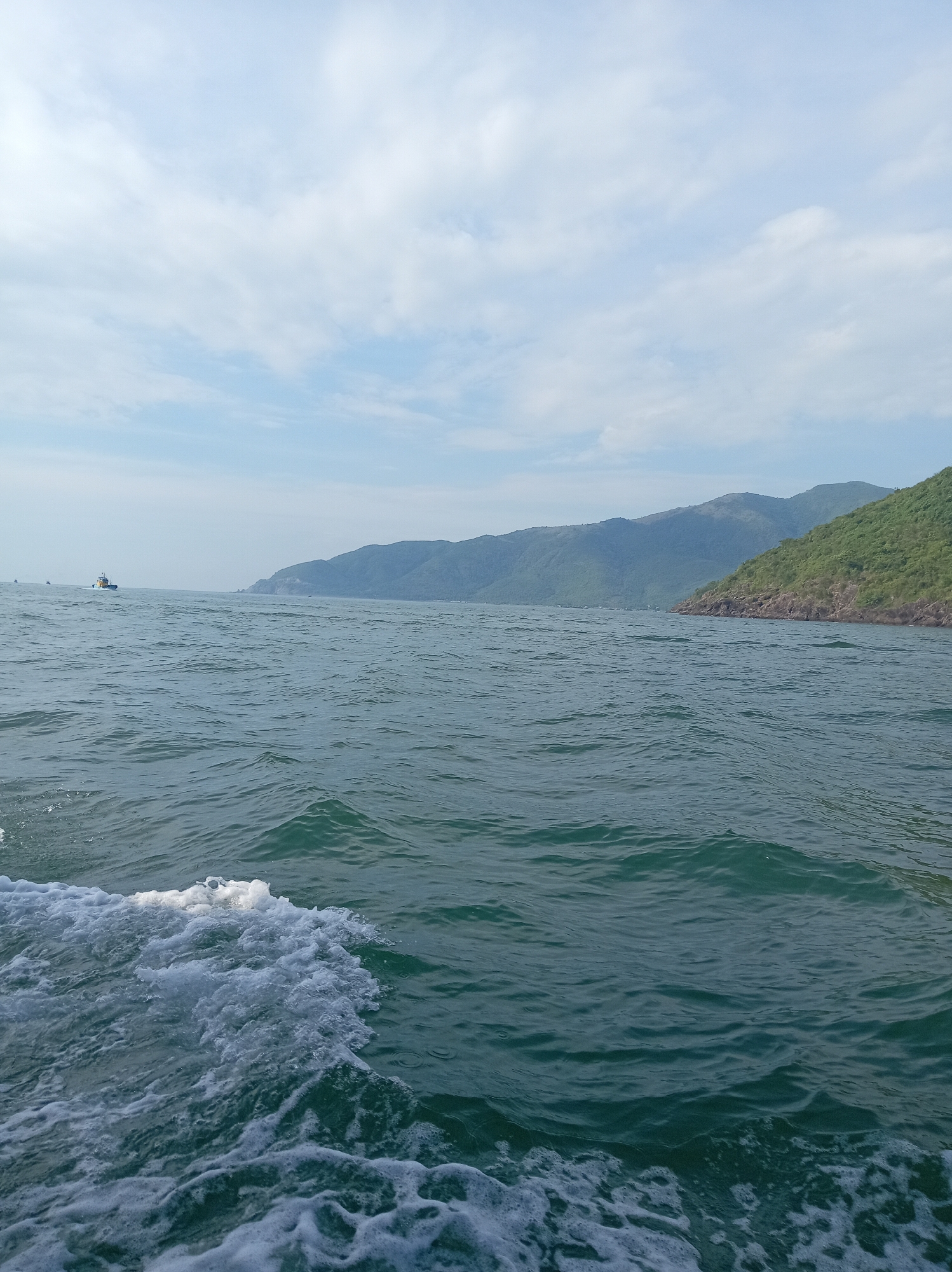 The image is a wide shot of the ocean, taken from a boat. The water is choppy, with white foam visible on the left side of the frame, indicating the boat is moving. In the distance, a long, green, mountainous coastline stretches across the horizon under a partly cloudy sky. A small boat is visible on the left side of the frame, further out at sea. The scene appears to be a scenic view of a coastal area.