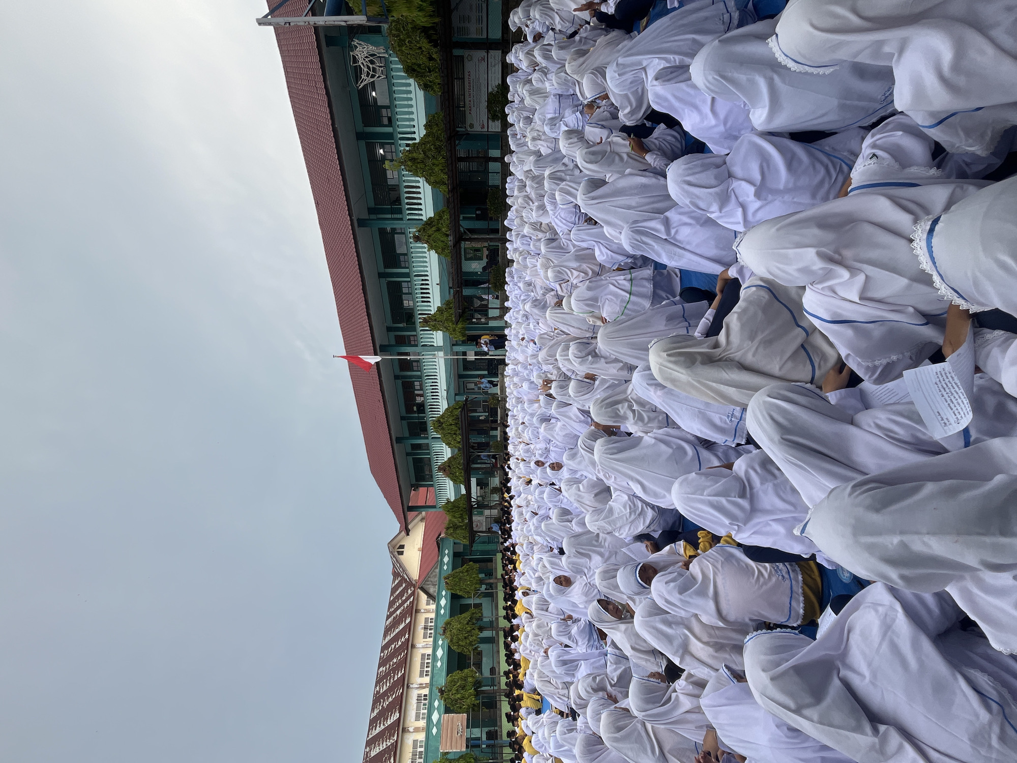 In Lhokseumawe, Indonesia, a large group of students, predominantly female, are gathered outdoors in what appears to be a school courtyard. They are all wearing white hijabs and some have blue accents on their clothing. The students are seated on the ground, facing towards a building in the background. The building has a red tiled roof and light green walls, with visible windows and balconies. An Indonesian flag flies on a flagpole in front of the building. Trees are planted along the front of the building. In the distance, other buildings are visible, including one with a striped roof. The sky is overcast, suggesting it might be a cloudy day. The overall scene depicts a school assembly or event.