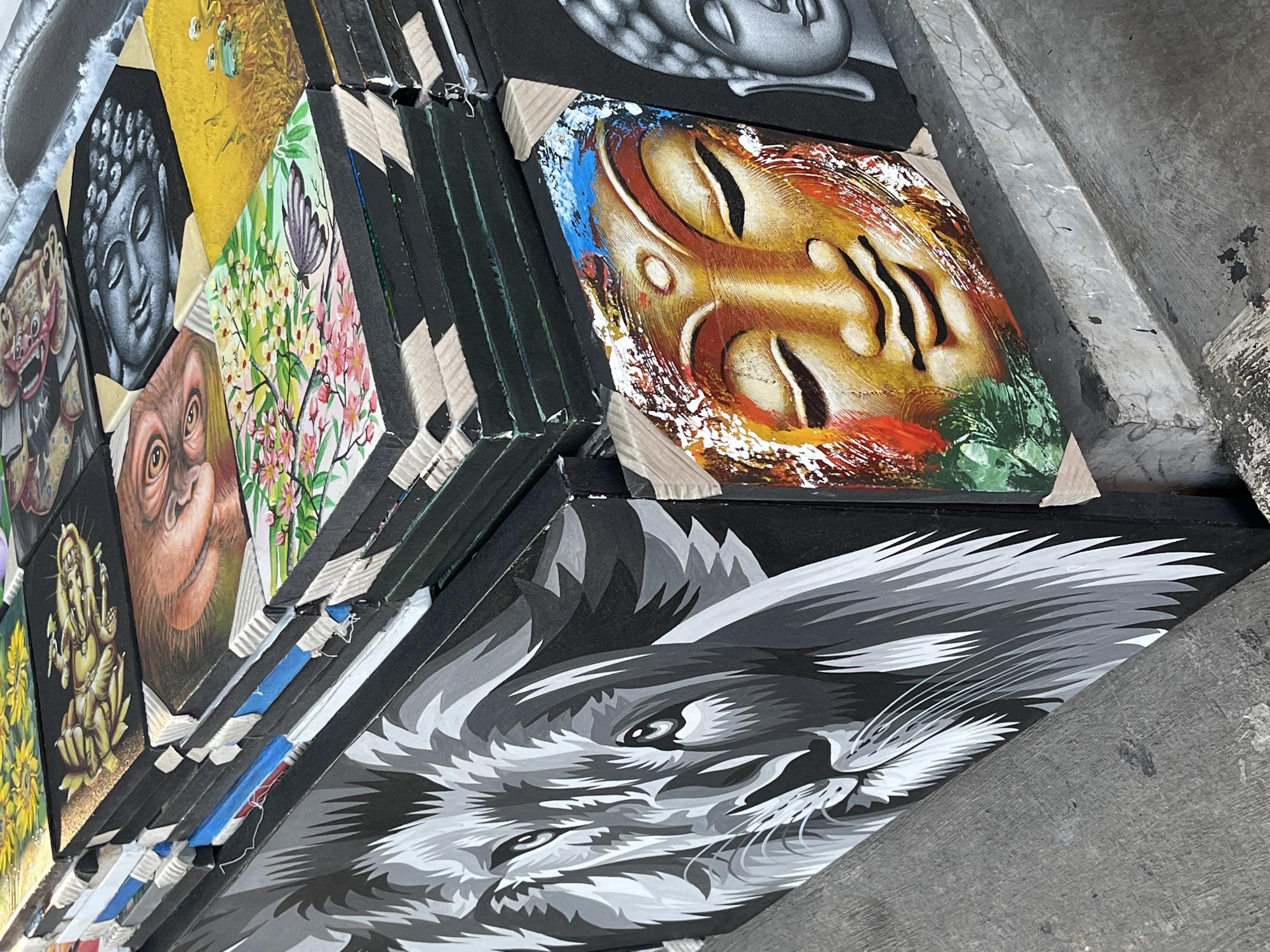 The image shows a stack of framed artworks in what appears to be an outdoor setting. The artworks include paintings of a wolf in grayscale, Buddha faces in various styles and colors, an orangutan, flowers, and a Balinese deity. The paintings are stacked on top of each other, with the frames visible along the edges. The environment looks like a street or sidewalk with a concrete surface. The location is in the city of Unknown, Indonesia.