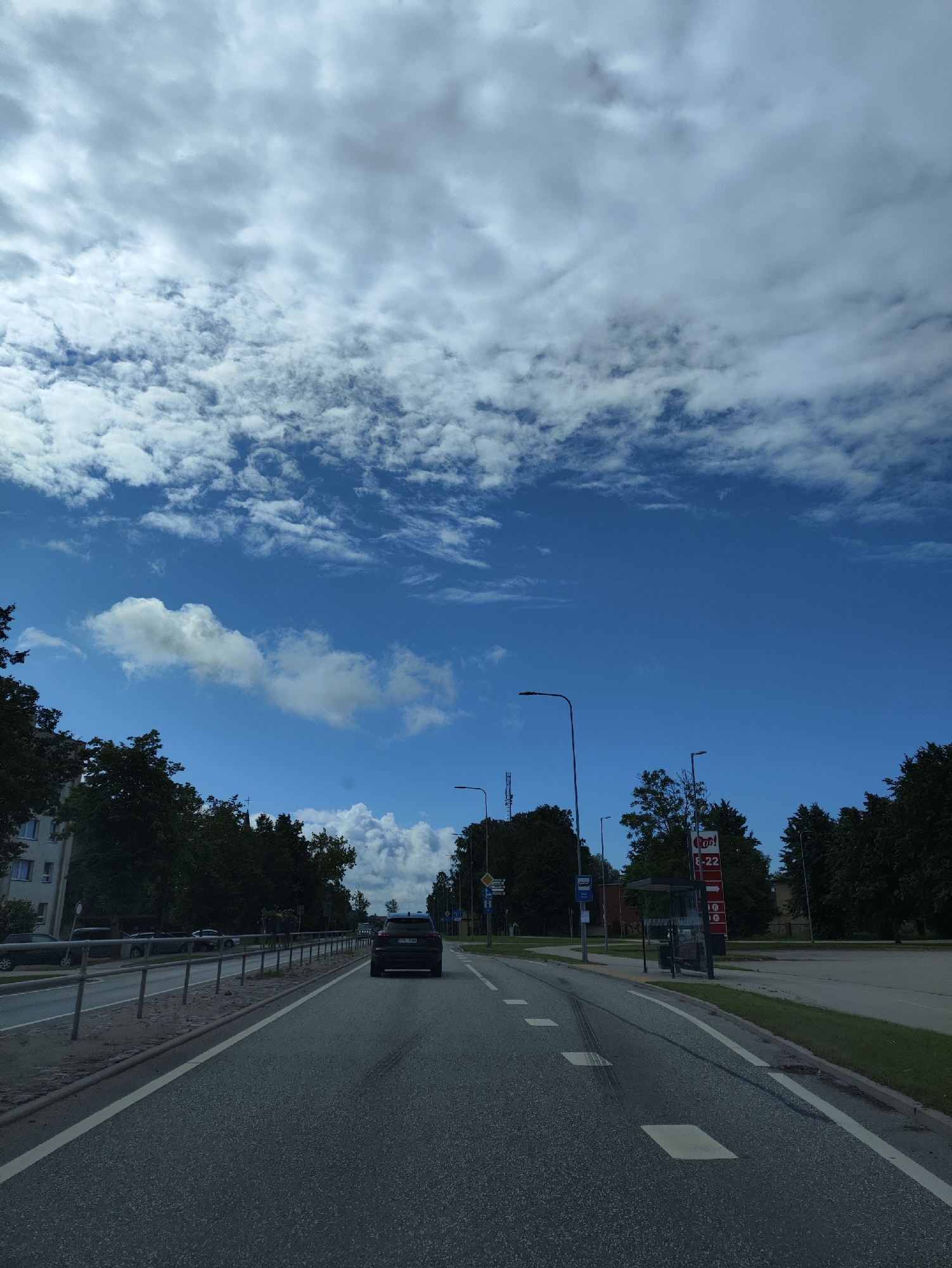 The image captures a street scene in Salacgrīva, Latvia, on a partially cloudy day. A dark-colored SUV is driving away from the camera on the right lane of a paved road. The road has white lane markings, including a solid white line on the right edge and dashed lines separating lanes. On the left side of the road, there is a metal fence separating the road from a sidewalk and a building. Trees and some parked cars are visible behind the fence. On the right side of the road, there are more trees, a bus stop shelter, and a red sign with white text that appears to be for a service station, displaying hours from 6-22. Streetlights are present on both sides of the road. The sky is a vibrant blue with scattered white and gray clouds.