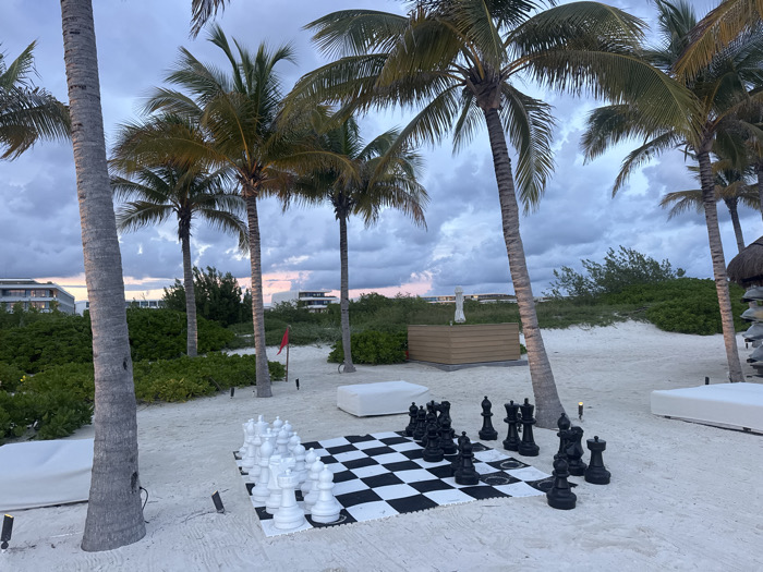 A beach scene at dusk features a giant chess set with white and black pieces arrayed on a large checkerboard mat. Palm trees frame the scene, and lush green foliage and a few buildings are visible in the background. A small white statue stands on a rectangular wooden platform. The sky is cloudy with hints of pink, suggesting either sunrise or sunset.