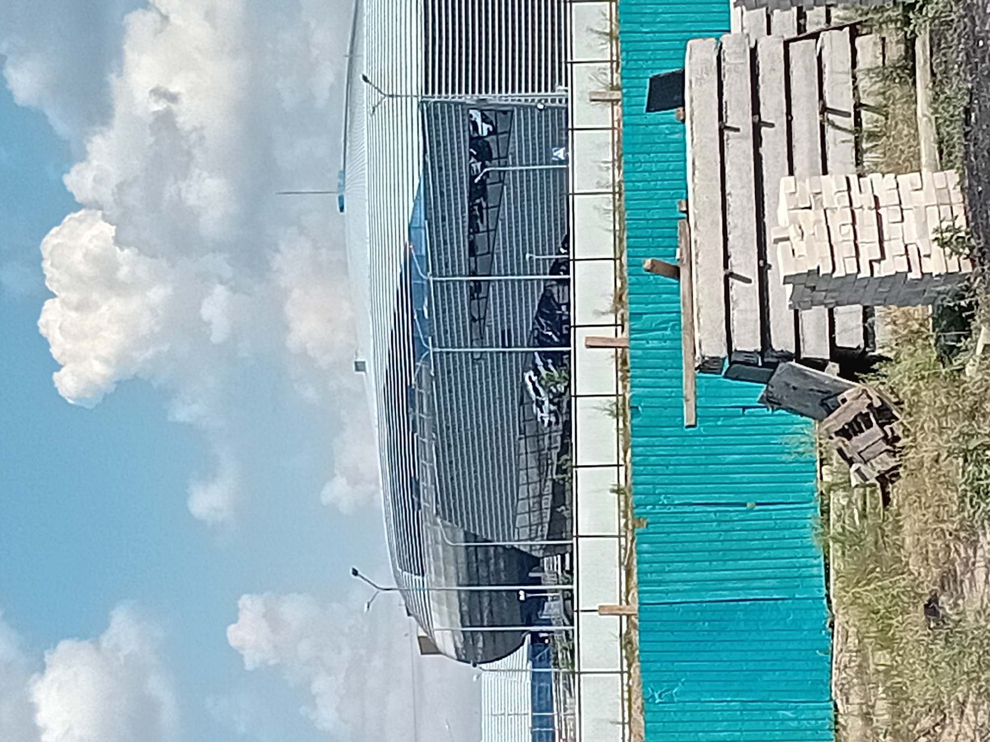 The image depicts an outdoor construction site in Semey, Kazakhstan, on a bright, cloudy day. In the background, a large, modern building with a distinctive curved, silver facade and horizontal slatted windows dominates the scene. The building appears to be a stadium or arena. In the foreground, a bright turquoise metal fence runs horizontally across the image, with a white picket fence visible behind it. Piled in front of the turquoise fence are stacks of concrete slabs and a smaller stack of light-colored bricks. The ground is covered in dry grass and dirt. No people are visible in the frame.