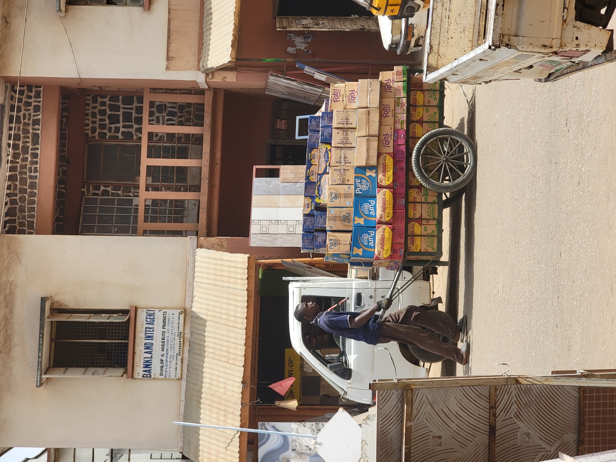 A man pulls a cart loaded with boxes on a paved street in Maiburiji, Nigeria. The cart, with a single wheel, is laden with numerous cardboard boxes containing various products, including "Pure Bliss", "ADAO" and "Fabi!" drinks, and tiles. The man is dressed in a dark blue shirt and dark trousers. He is pulling the cart towards the right of the frame. A white van and a three-wheeled vehicle are parked nearby. Buildings line the street, including one with a sign reading "BANKLAND INTER AGENCY," advertising Dunlop and Nigerite products. The buildings are made from concrete and are painted a beige color.