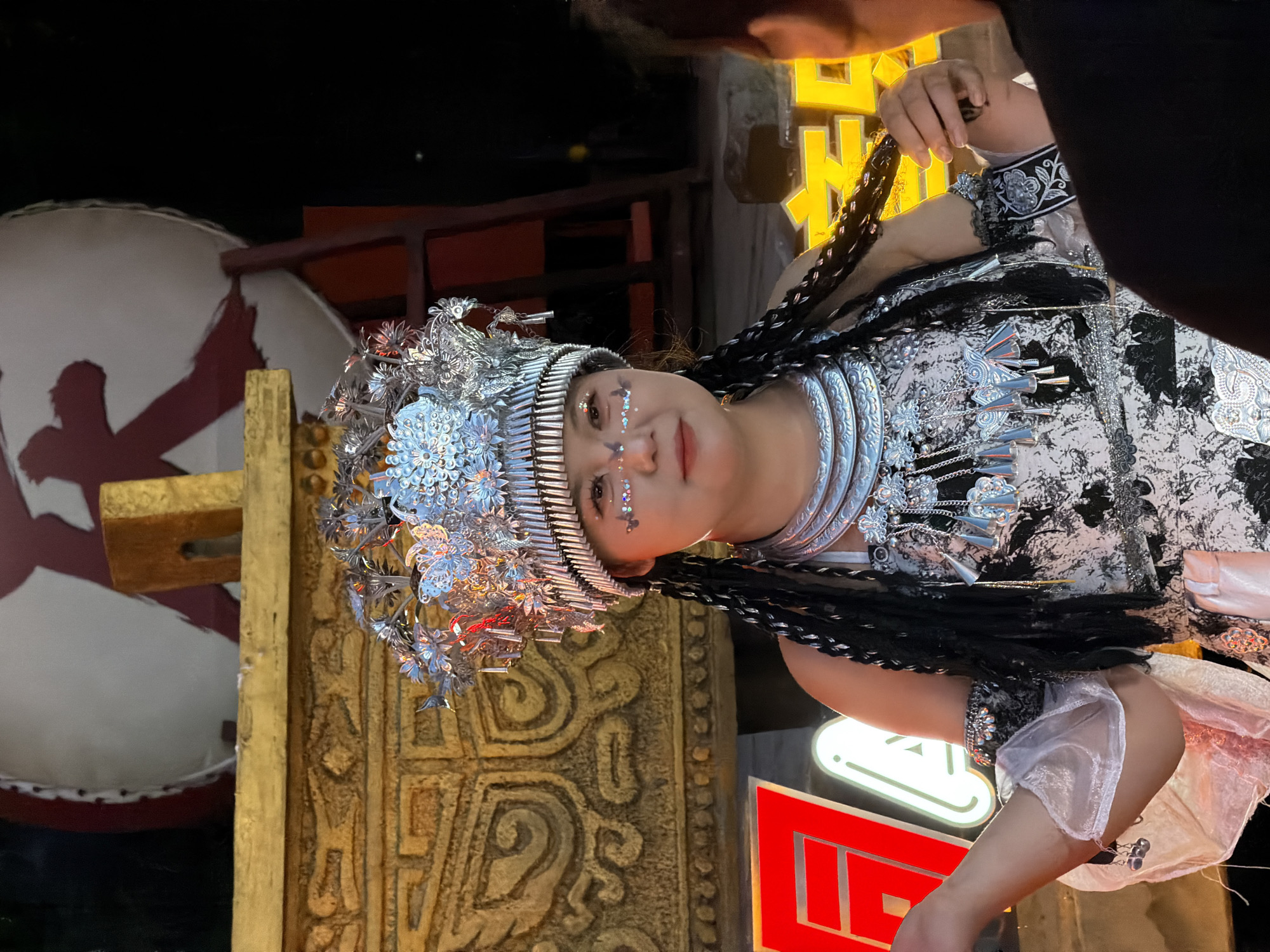 A person in traditional dress, adorned with intricate silver headwear and jewelry, is the focus of this image. The costume features a patterned garment and flowing sleeves. The individual's face is lightly decorated with glitter on the cheeks and eyelids, and they have a serene expression. Their dark hair is styled in braids that hang down.
The scene appears to be outdoors at night, with artificial lighting illuminating the subject. Behind the person, there are some cultural decorative elements. To the left, a large, round object with a red design is partially visible, and a wooden structure with carved patterns is prominent. To the right, neon signs with Chinese characters are lit, suggesting an urban or commercial setting. The overall atmosphere is festive and perhaps ceremonial. The specific location is hinted at by the cultural attire and the presence of Chinese characters on the signs, which aligns with the provided context of Yuzhong District, China.
