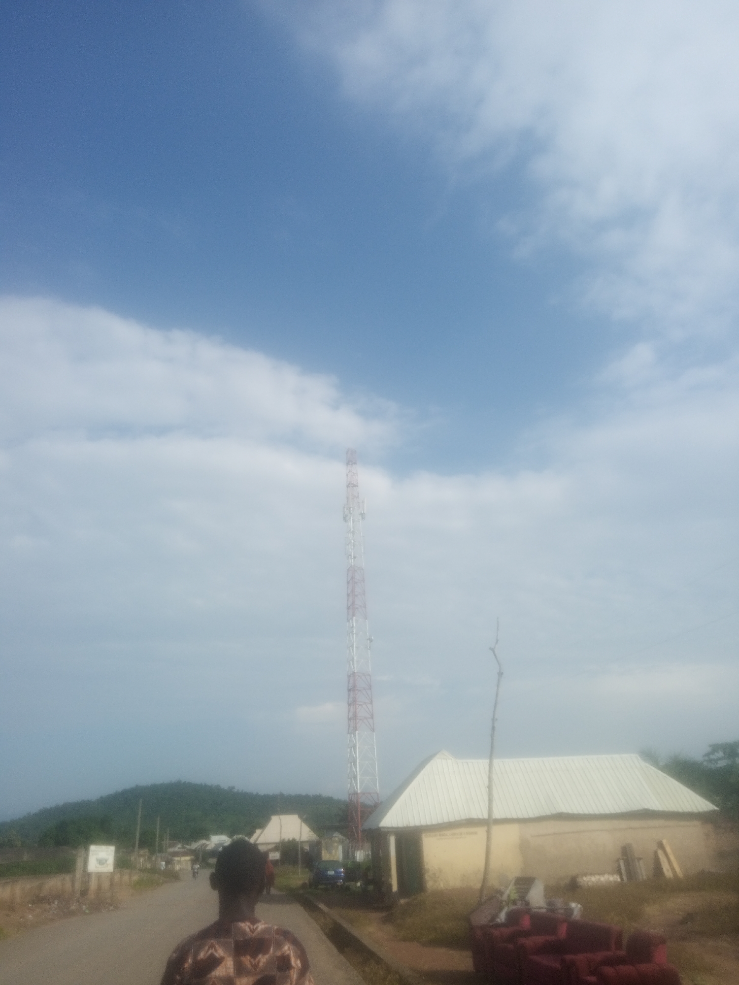 This image depicts an outdoor scene in Serti, Nigeria. In the foreground, the back of a person wearing a patterned shirt is visible. The scene is a street with a telecommunications tower on the right side. Further down the road, there are several buildings, including a light-colored structure with a white metal roof, and a distant green hill. The sky is partly cloudy with a clear blue sky above the clouds. The lighting suggests it is daytime, possibly mid-morning or afternoon, with diffuse light due to the cloud cover. There are no explicit signs of a specific event taking place, appearing to be a typical day in a semi-rural setting. The overall mood is neutral, reflecting a quiet, everyday moment. No discernible text is visible in the image.
