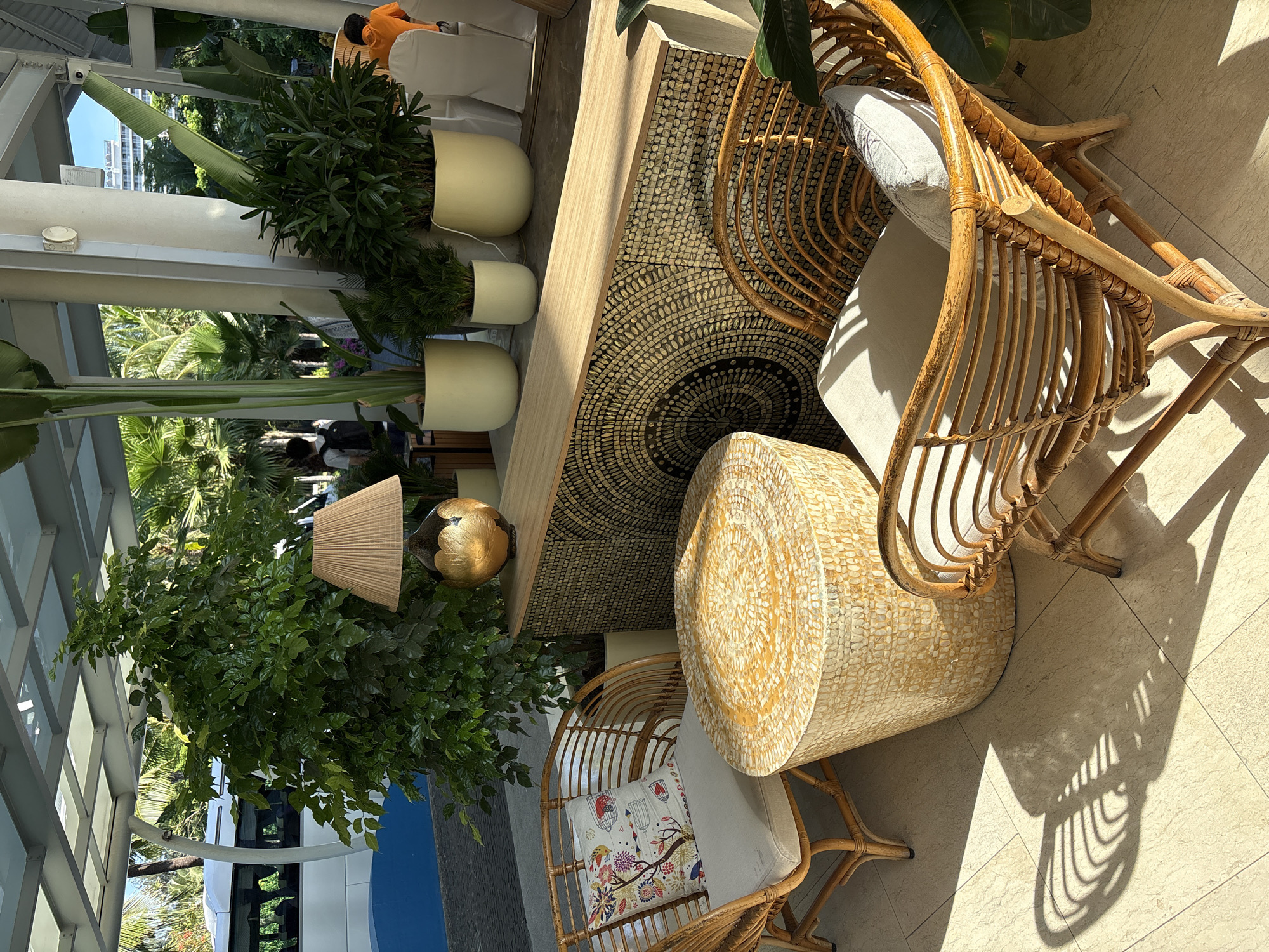 The image shows an outdoor seating area at a resort, likely in the city of Đà Nẵng, Vietnam, given the tropical foliage and architecture. Two wicker chairs with light-colored cushions and decorative pillows are arranged around a cylindrical, textured table. A tall, lush green plant stands behind the chairs, and several other potted plants are visible in the background. A long, narrow credenza with a patterned front and a light wood top is situated behind the table and chairs, and a lamp with a beige shade and a gold base sits on top of it. Sunlight casts strong shadows on the tiled floor. In the background, a white bus and palm trees are visible, suggesting a resort or hotel entrance. One person in an orange shirt is partially visible on the right side of the frame.