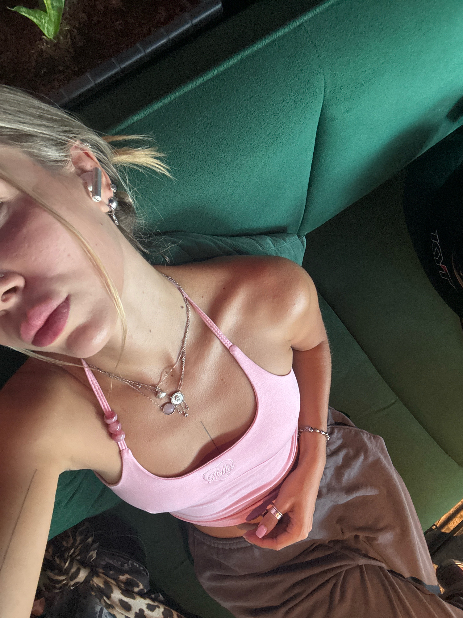 An overhead shot captures a young woman lying on a green sofa. Her hair is blonde and pulled back. She wears a pink tank top, gray sweatpants, and several necklaces and rings. One necklace has a circular pendant with a shell and pink stone. Her left ear has multiple piercings. To her right, a black object with white lettering is visible. The background is a dark, textured surface, possibly a plant pot. The lighting suggests a natural light source, casting shadows.